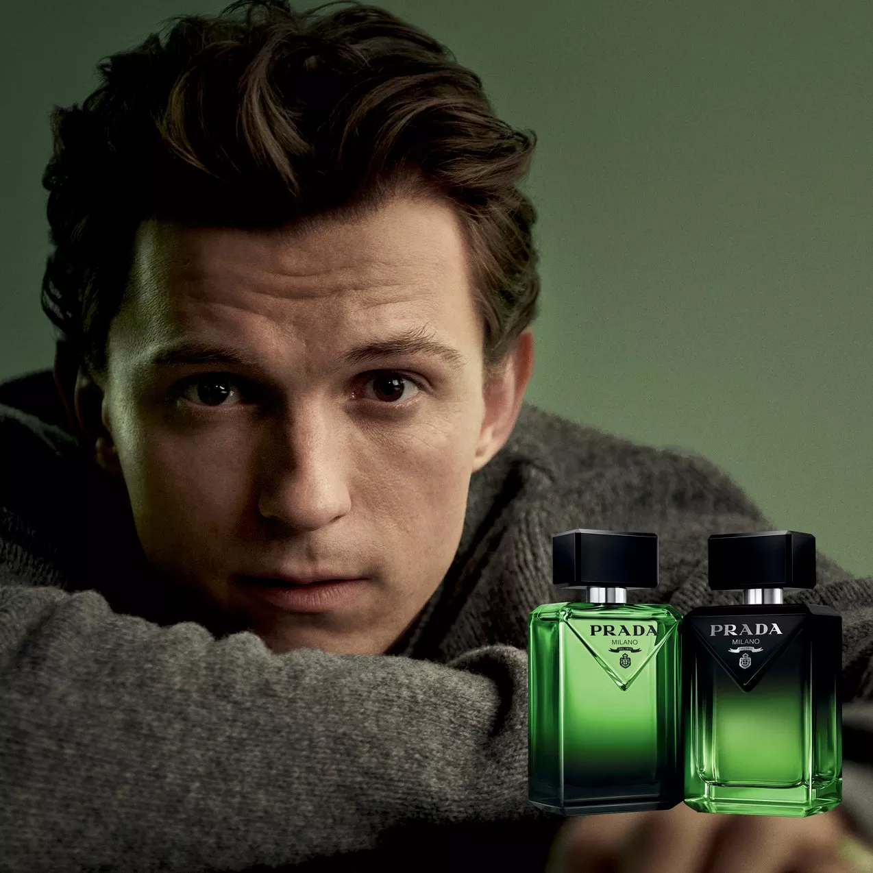 Actor Tom Holland resting his chin on his arms beside two green Prada Milano fragrance bottles against a muted green background.