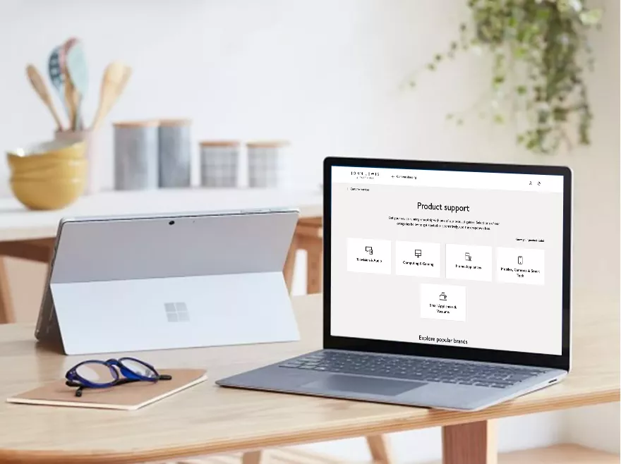 Two laptops on a wooden desk in a bright home office, one showing Windows 10 Start menu and apps.