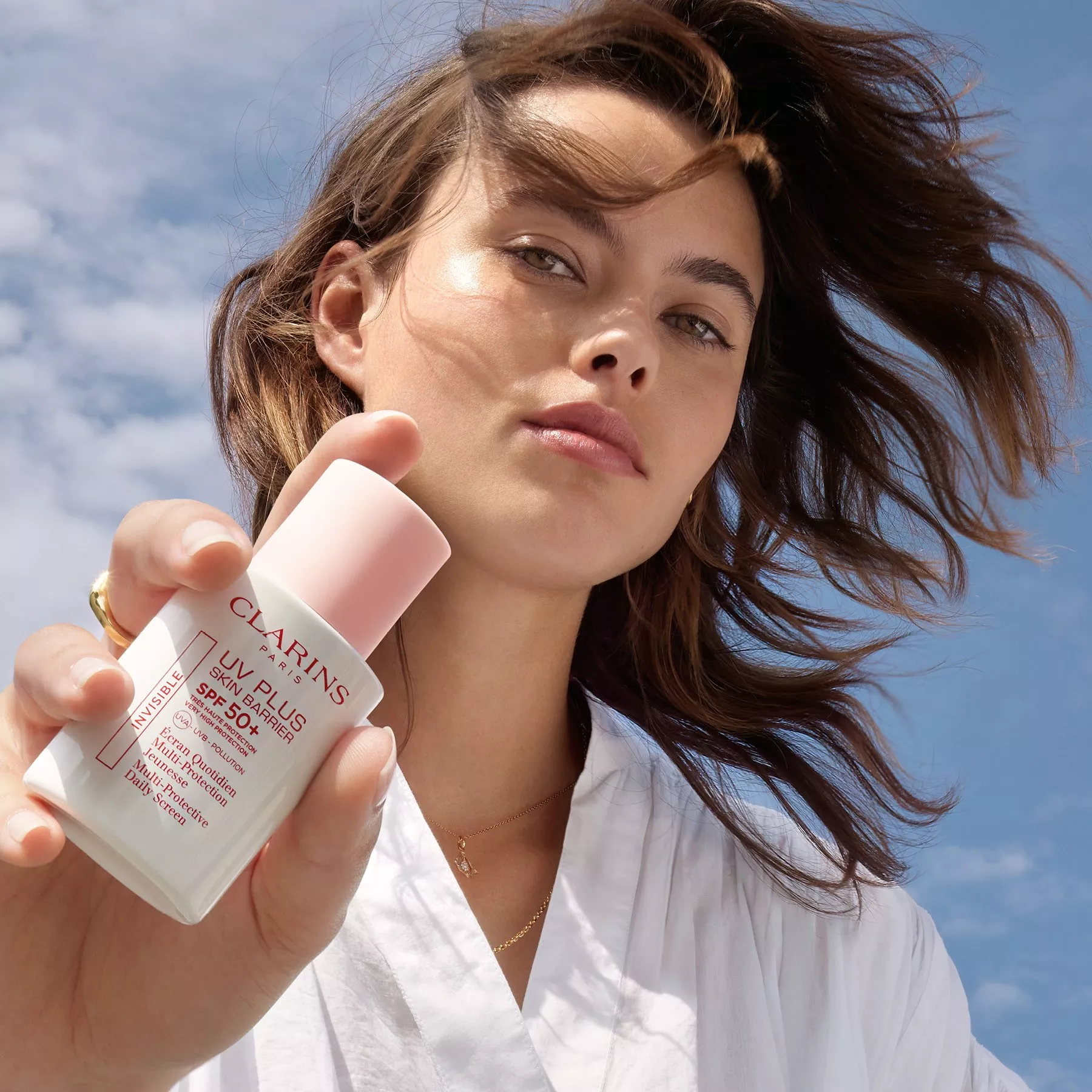 Woman holding Clarins UV Plus SPF50+ daily sunscreen, close-up skincare product shot against blue sky