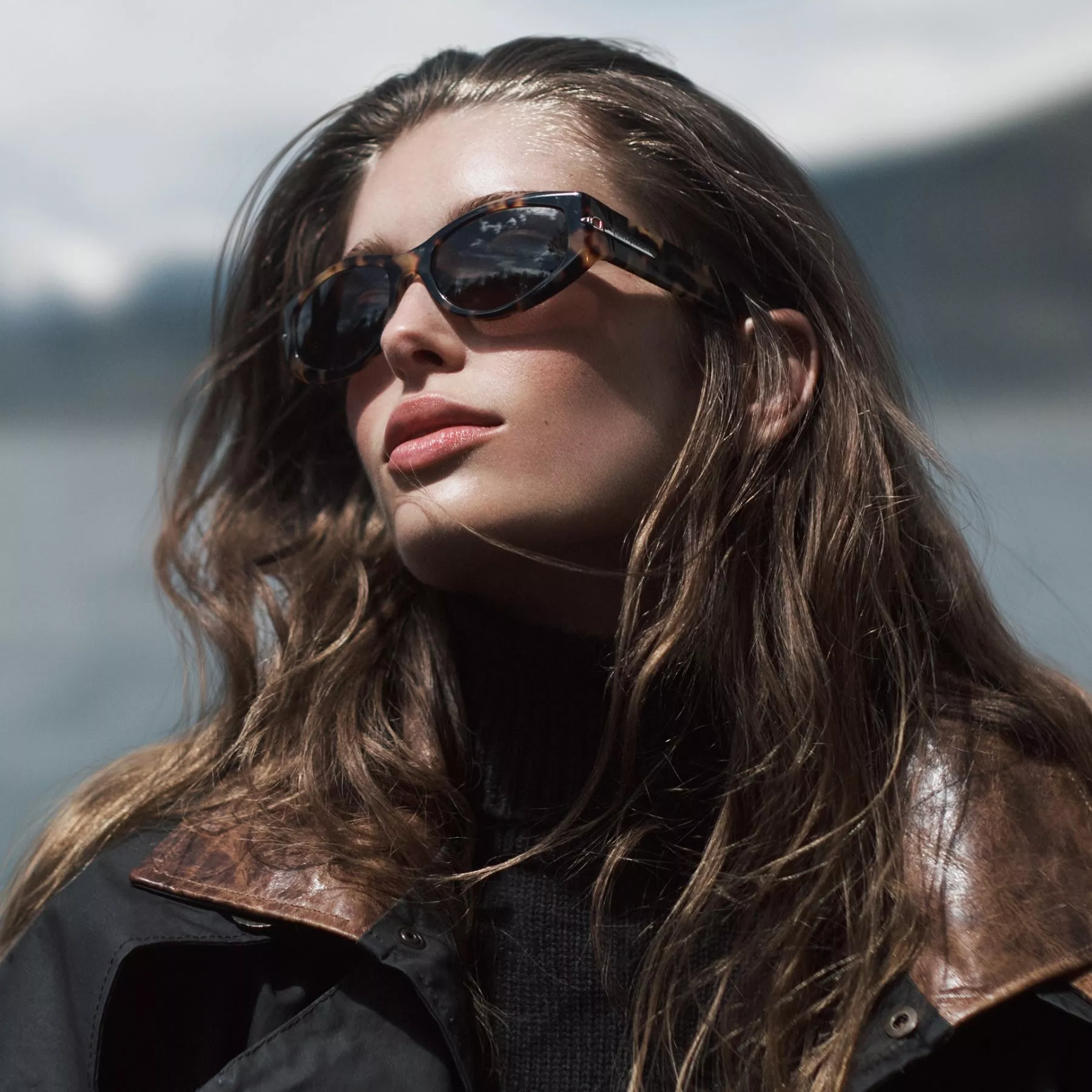 Stylish woman in sunglasses with brown leather shoulder bag by paddock fence, horses grazing at sunset
