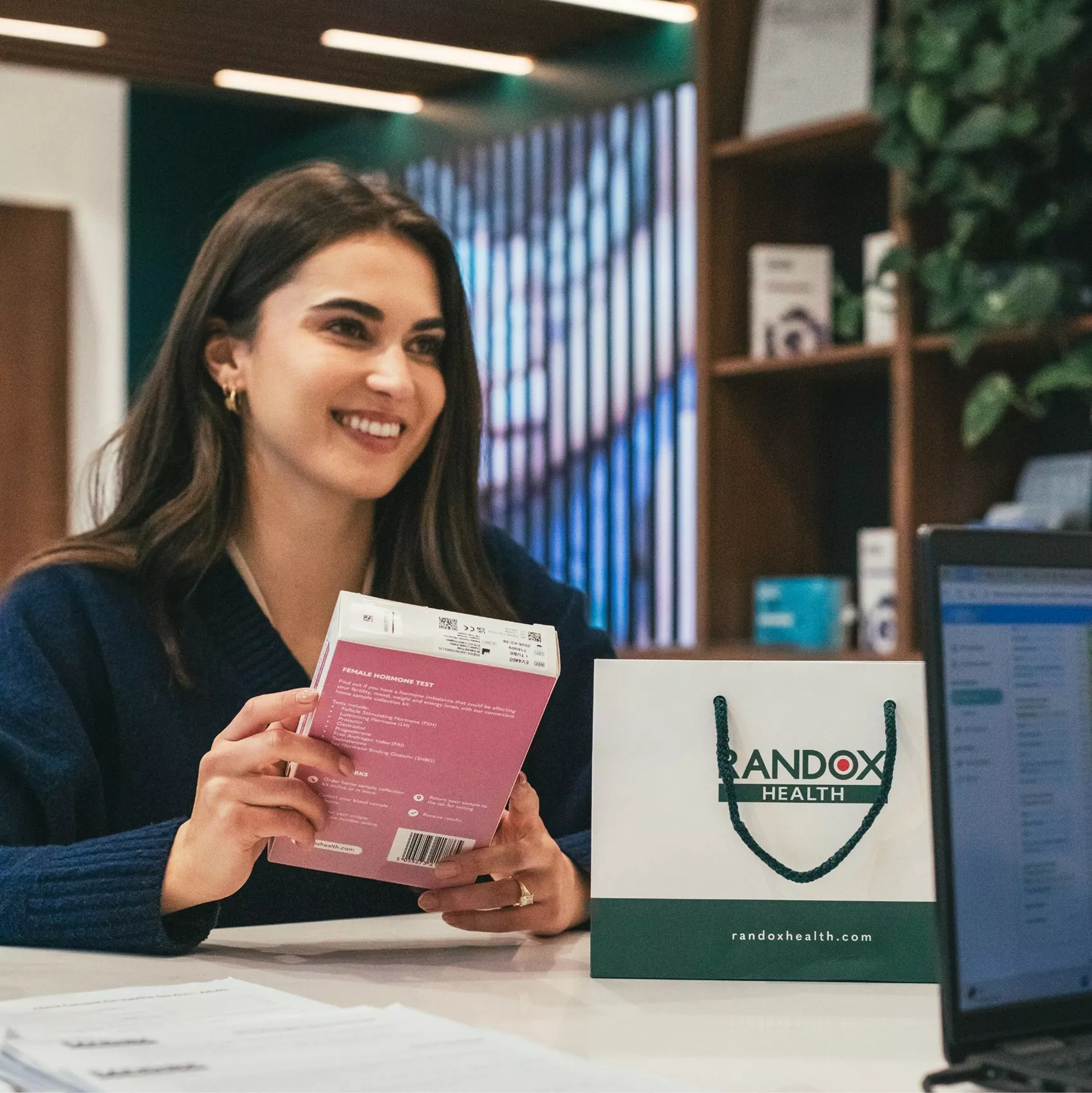 Smiling woman holding Randox Health female hormone test kit beside branded bag in modern clinic reception area