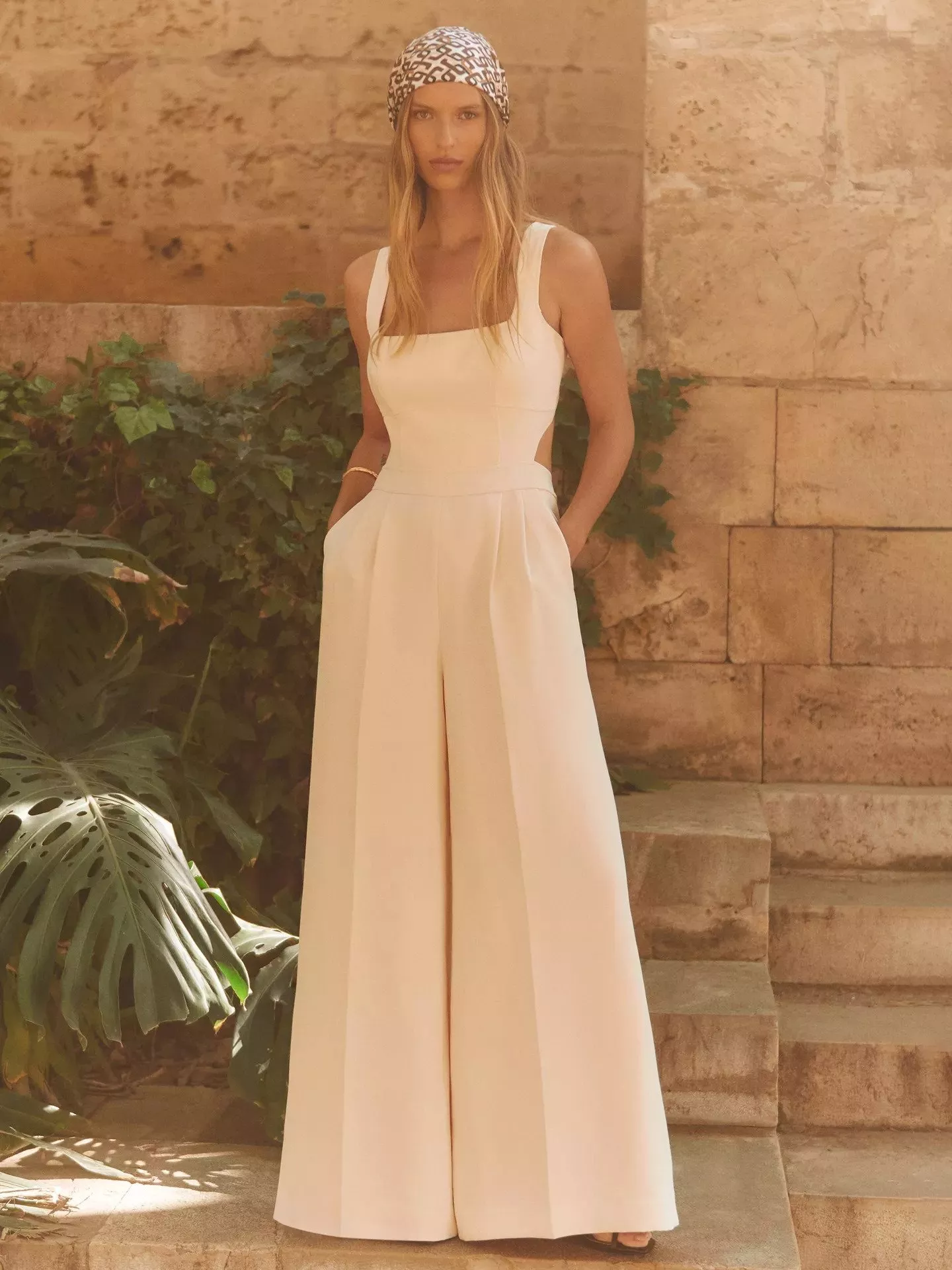 Woman in a beige jumpsuit and patterned headscarf stands on stone steps with leafy plants around her.