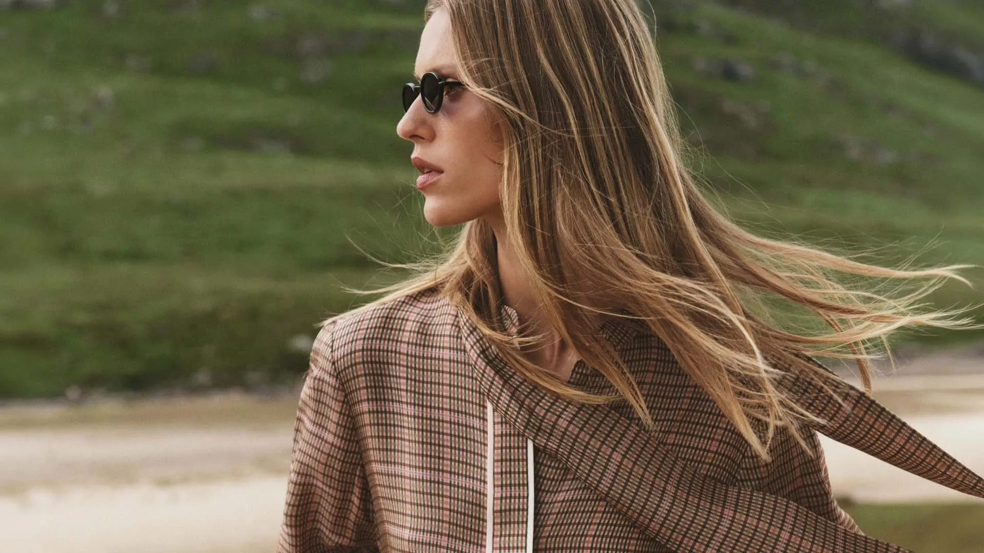 Profile view of a long-haired blonde woman wearing round dark sunglasses and a plaid top with a draped scarf, with her hair blowing in the wind against a grassy hill background.