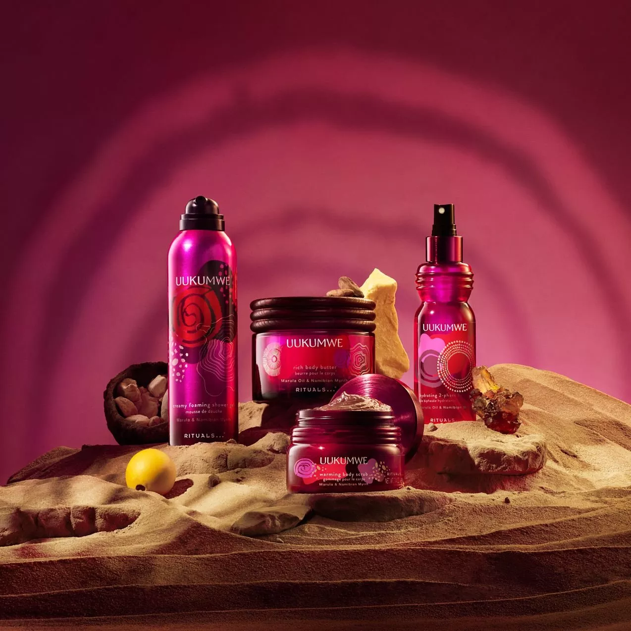 Rituals Uukumwe body care products—foaming shower gel, body butter, body scrub, and spray—displayed on sand against a pink-lit backdrop with raw natural ingredients.