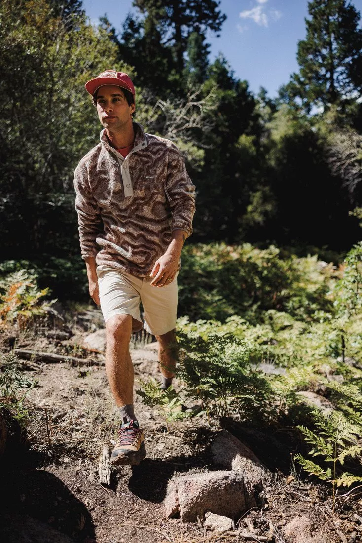 Man hiking on rocky forest trail wearing patterned fleece and red cap, outdoor adventure lifestyle in woodland scenery