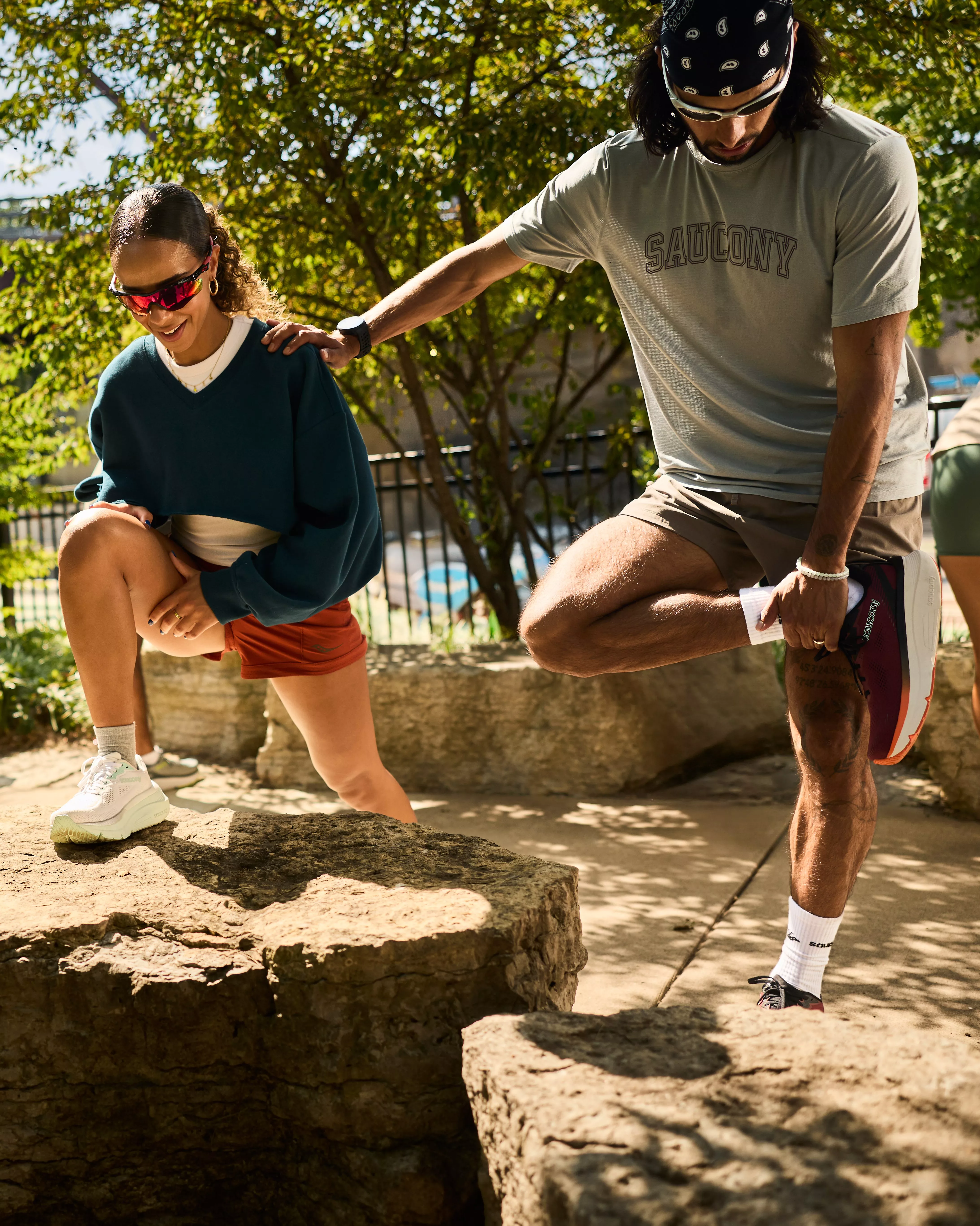 Two runners stretching outdoors in a sunny park, warming up for a run in trainers and sportswear