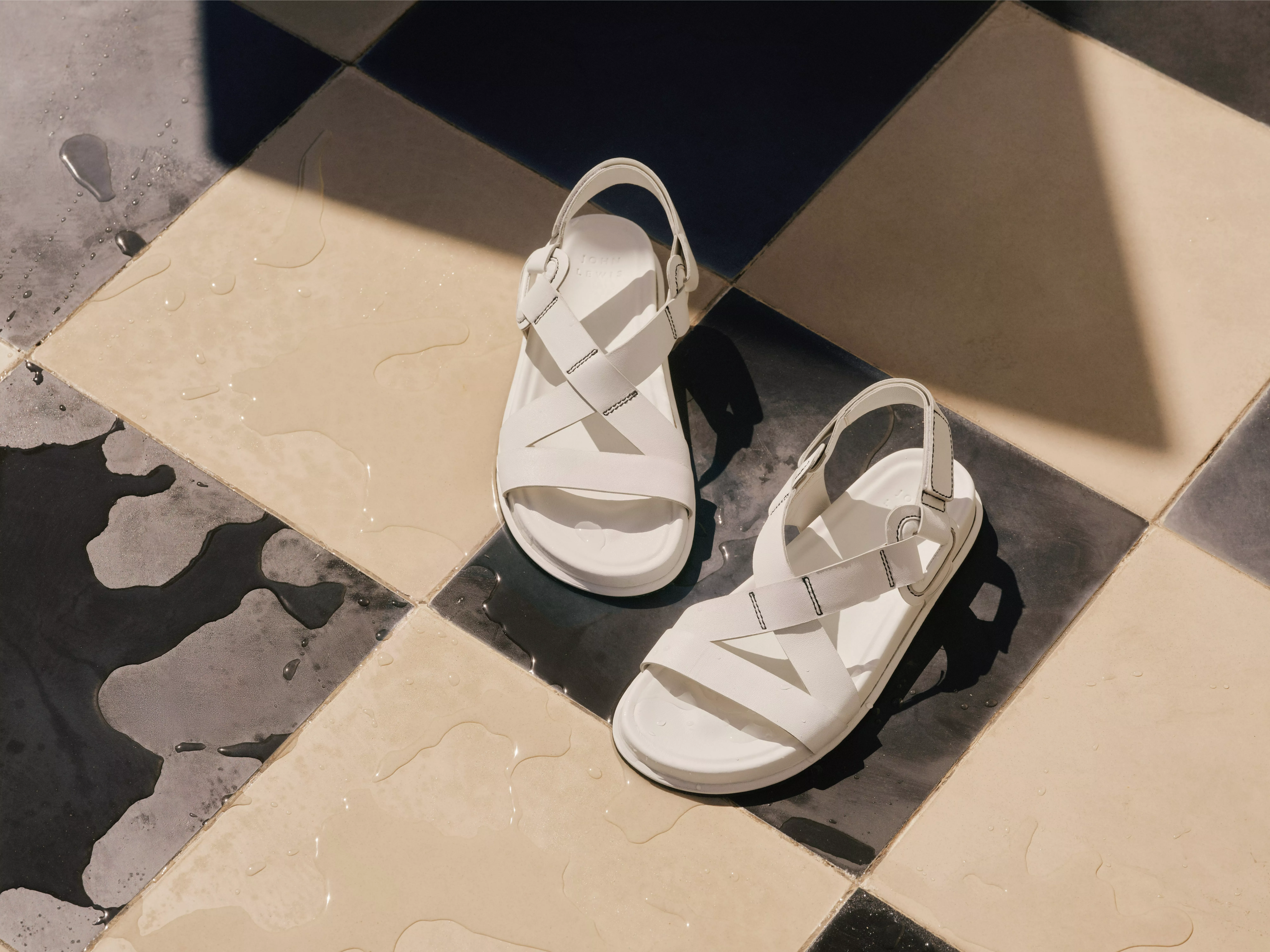 White leather sandals on wet black and cream tiled floor in sunlight, minimalist summer footwear product shot