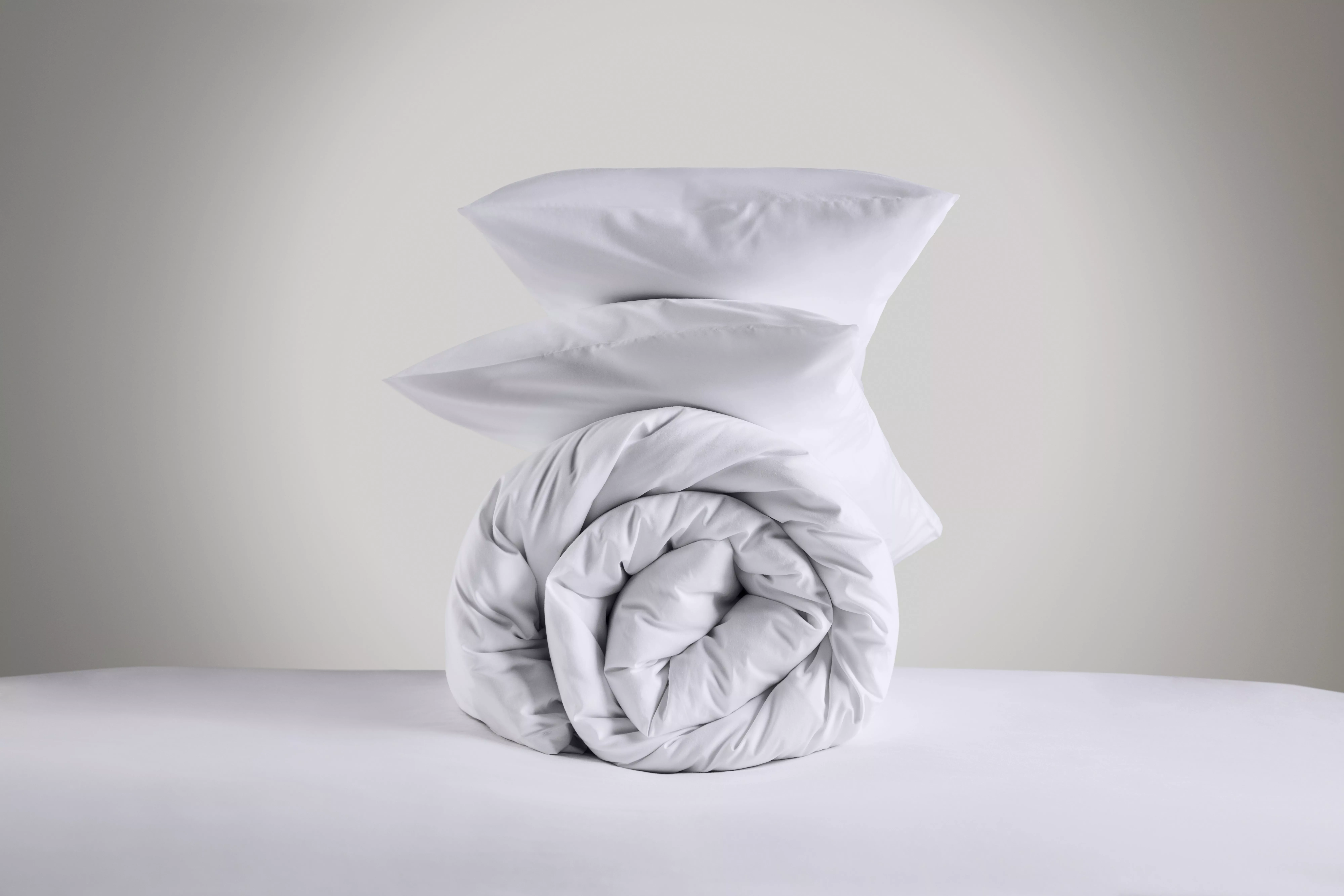 White bedding set with two pillows stacked on a rolled duvet, minimalist bedroom linen product photo
