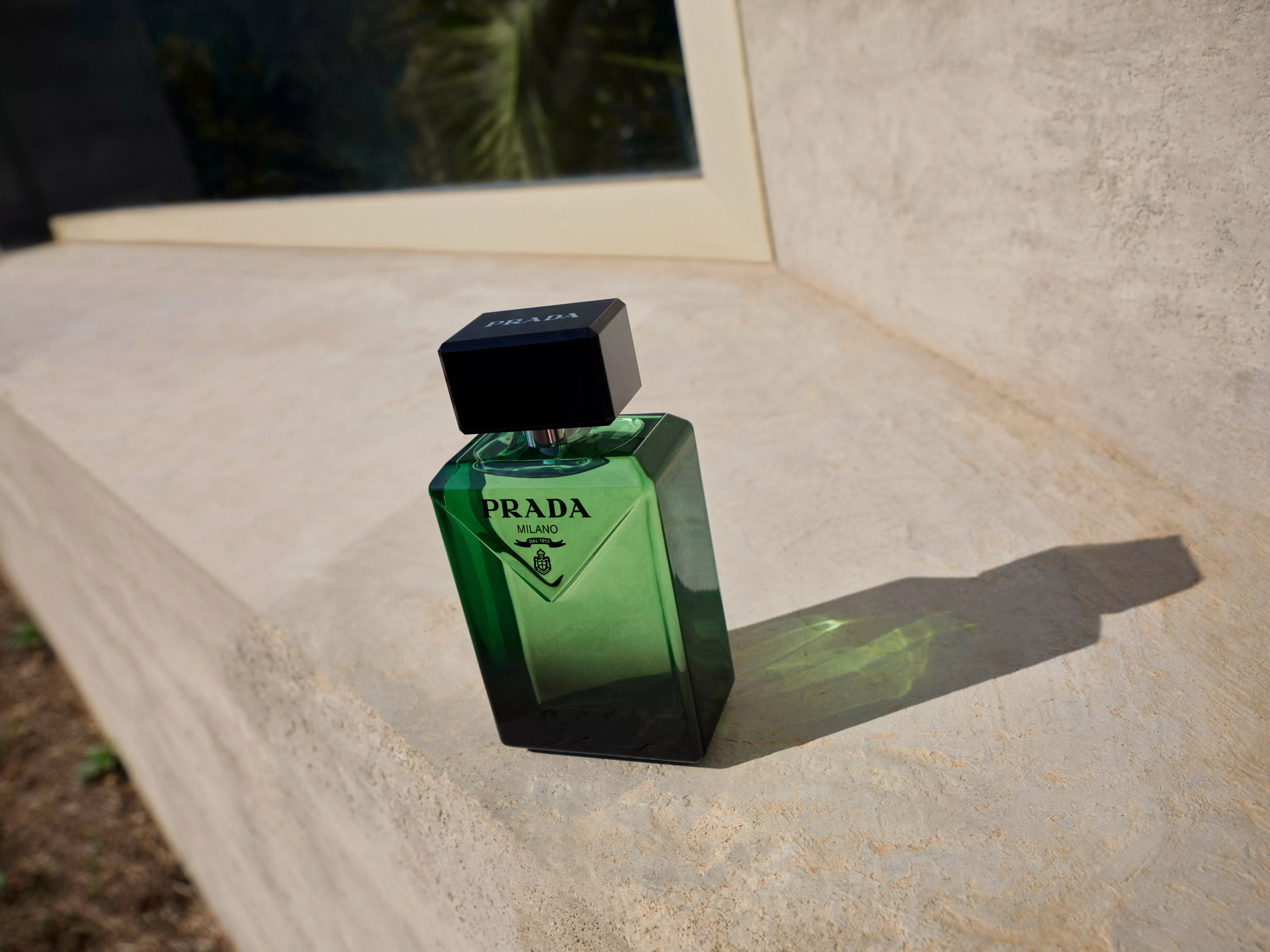 A green Prada perfume bottle with a black cap sits on a sunlit stone ledge, casting a shadow.