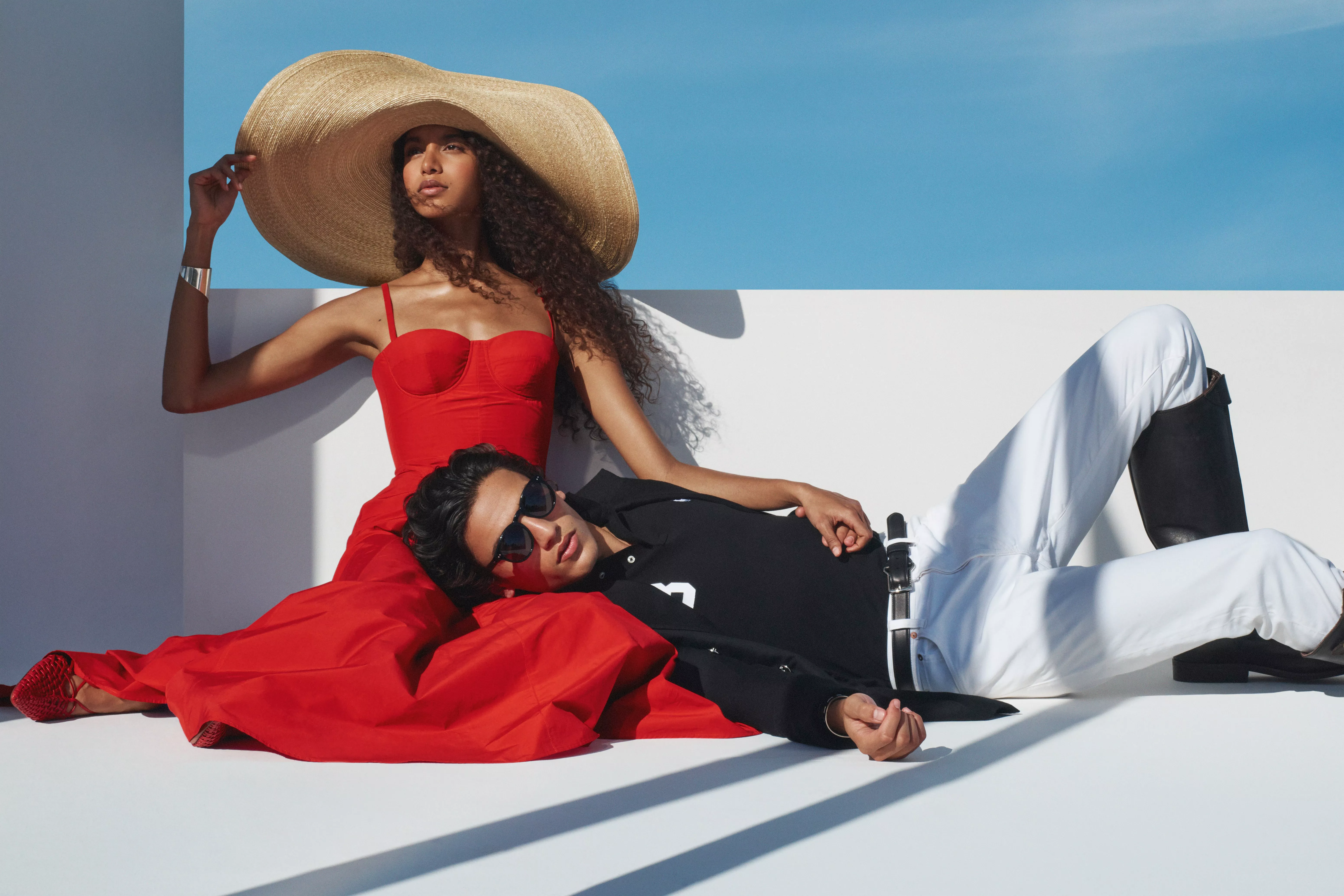 Fashion editorial of couple in resortwear: woman in red dress with wide-brim hat, man in black top and white trousers