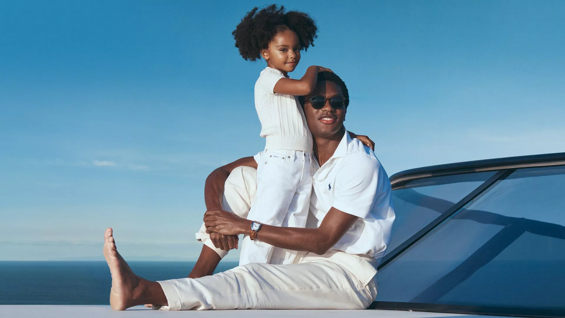 Stylish father in sunglasses with daughter in white outfits, seated by a luxury car with ocean backdrop.