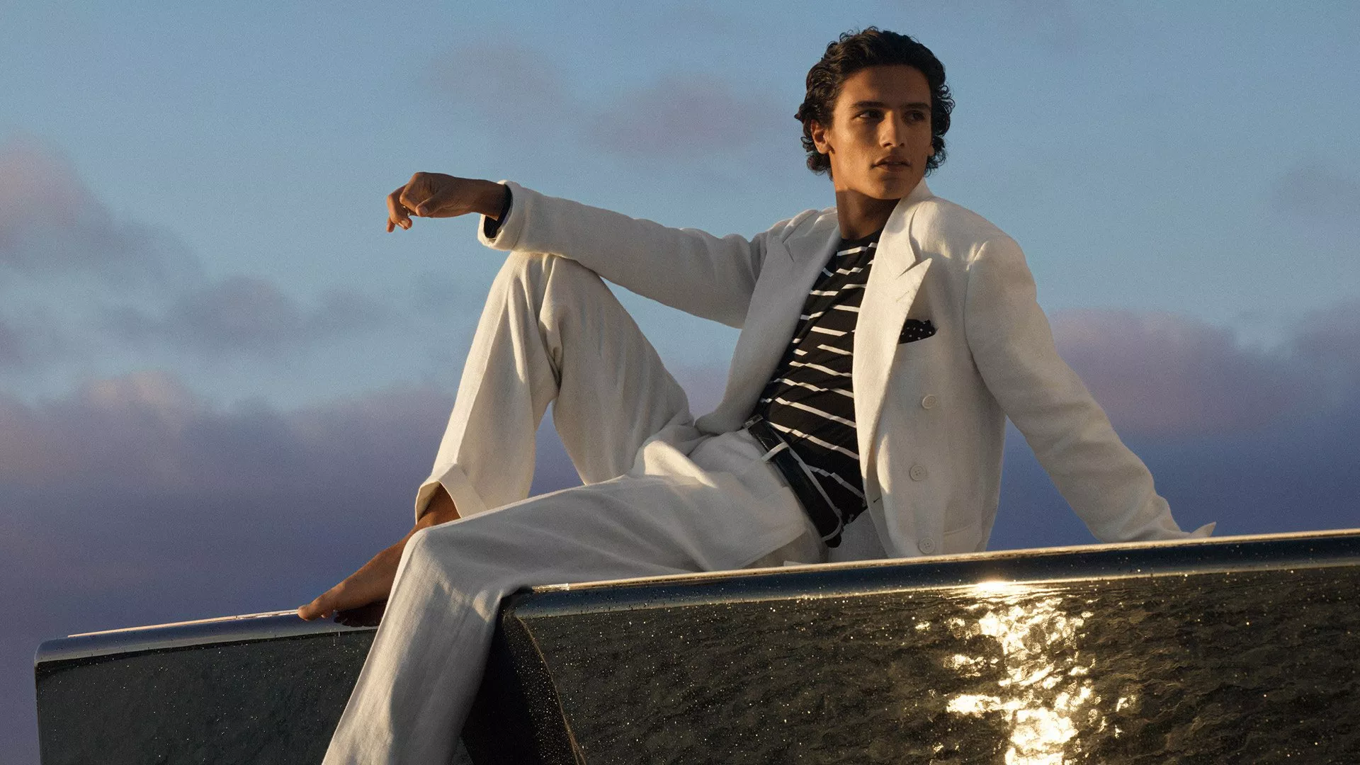 Stylish man in white suit and striped shirt posing on a yacht at sunset, luxury menswear fashion shoot