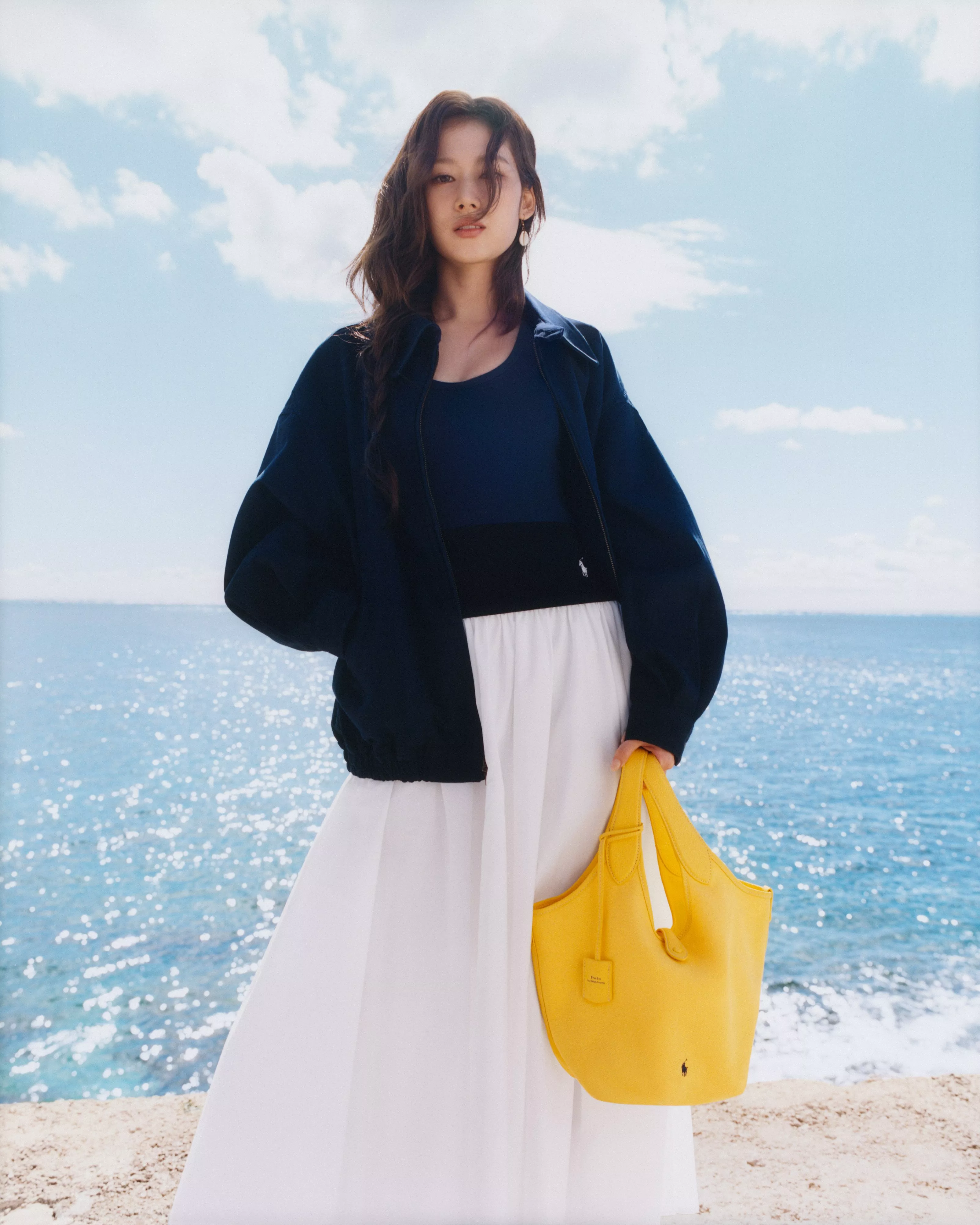 A woman in a black dress with gold buttons stands by a blue wall, holding a burgundy handbag, with saddles nearby.