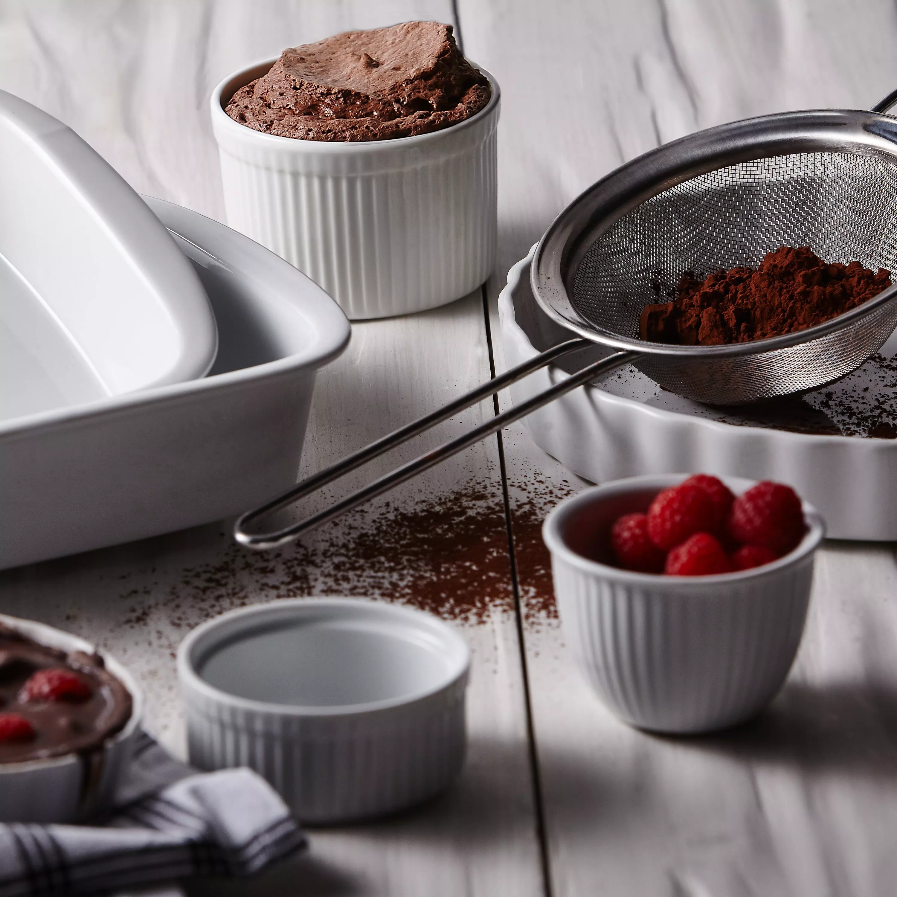 Chocolate soufflé in white ramekin with cocoa powder sieve, baking dish and fresh raspberries on table