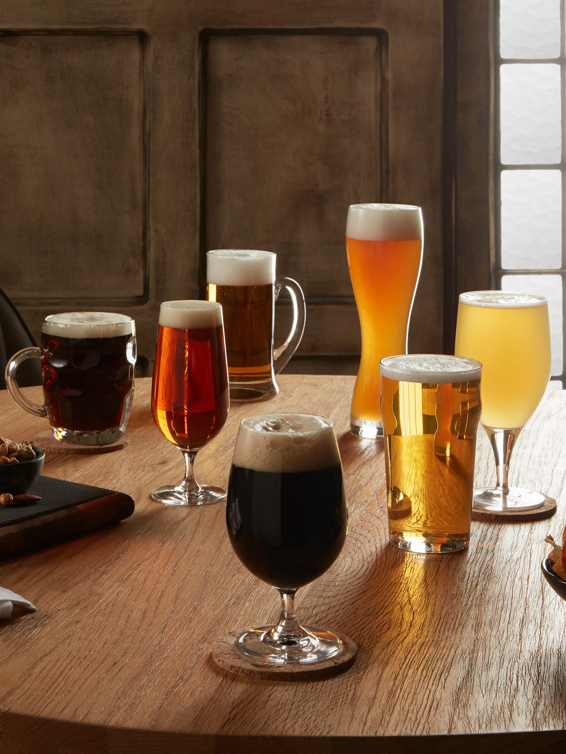 Craft beer bottle and four foamy beer glasses on rustic table with sliced salami and herbs, teal backdrop