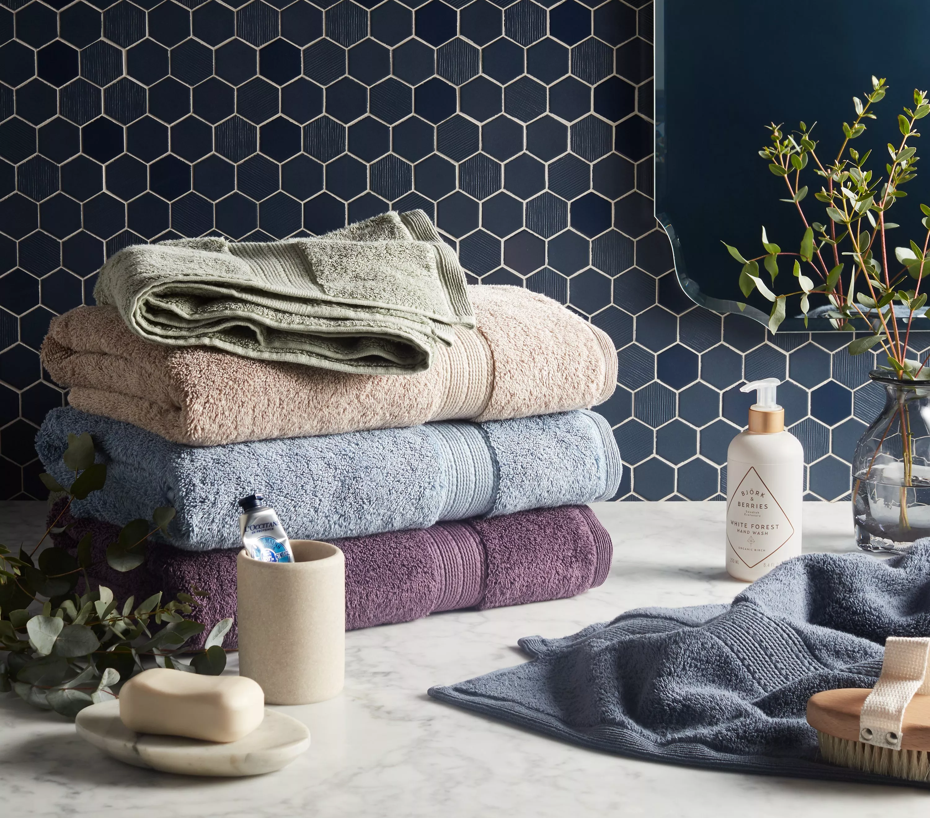 Stack of soft bath towels on marble counter with navy hexagon tiles, hand wash bottle and greenery in bathroom.
