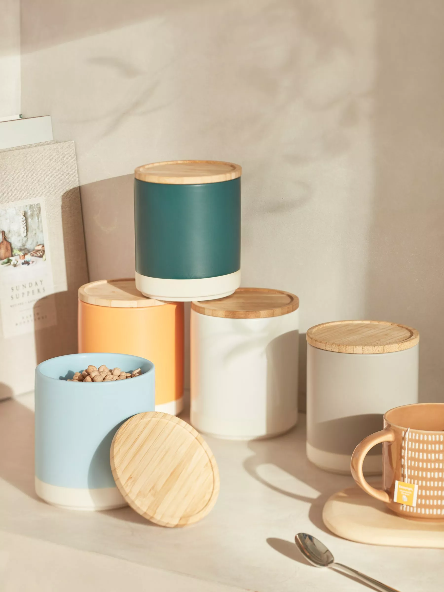 Colourful kitchen storage canisters with bamboo lids on countertop, plus mug and spoon in warm sunlight