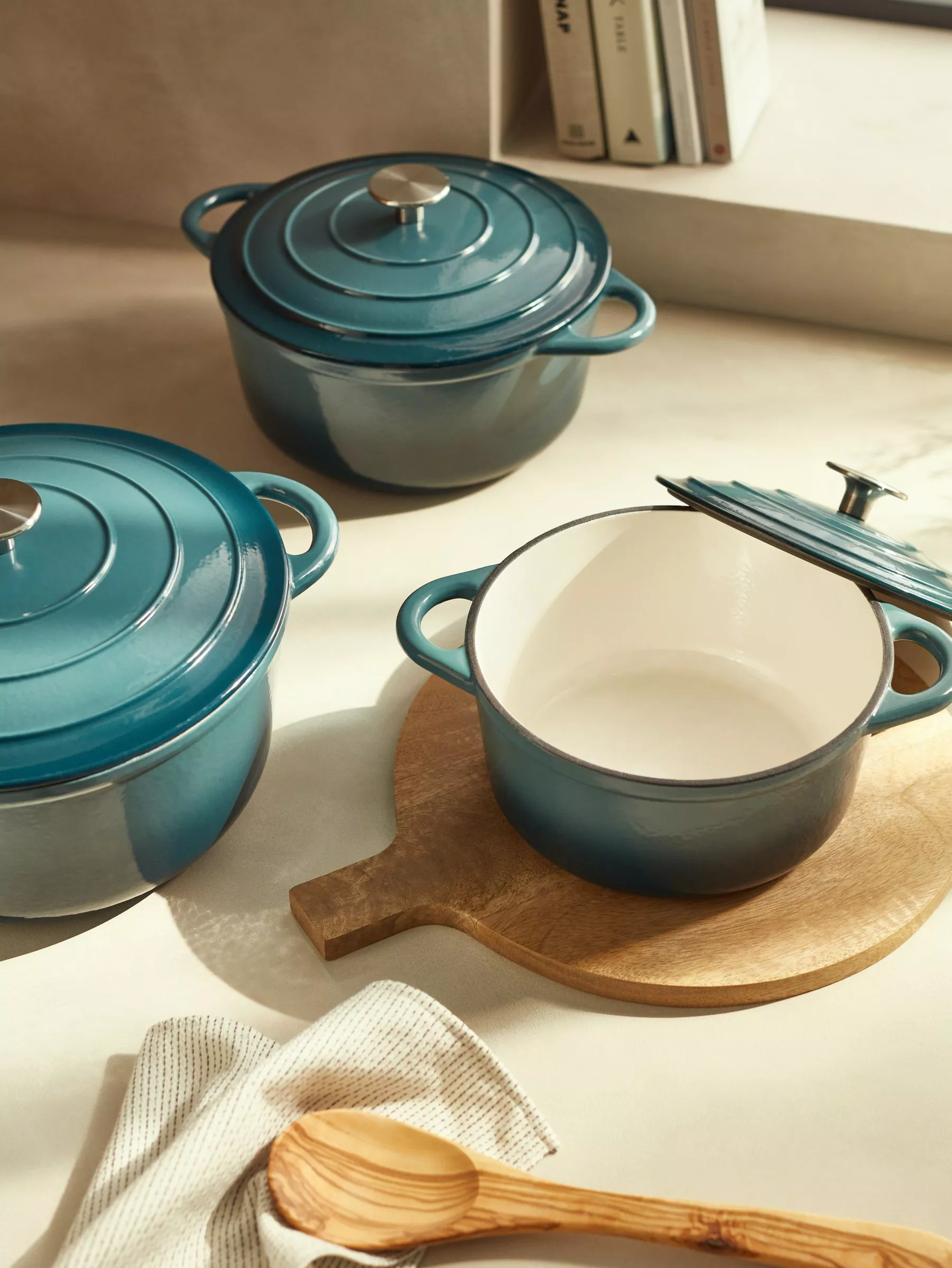 Teal enamel cast iron casserole dishes with lids on a kitchen worktop, one open on a wooden board