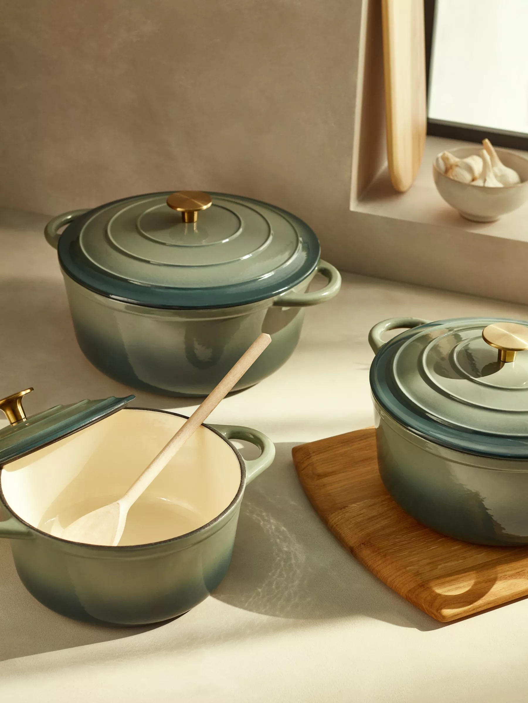 Green enamel cast iron casserole dishes and saucepan with lids, wooden spoon and chopping board in kitchen setting