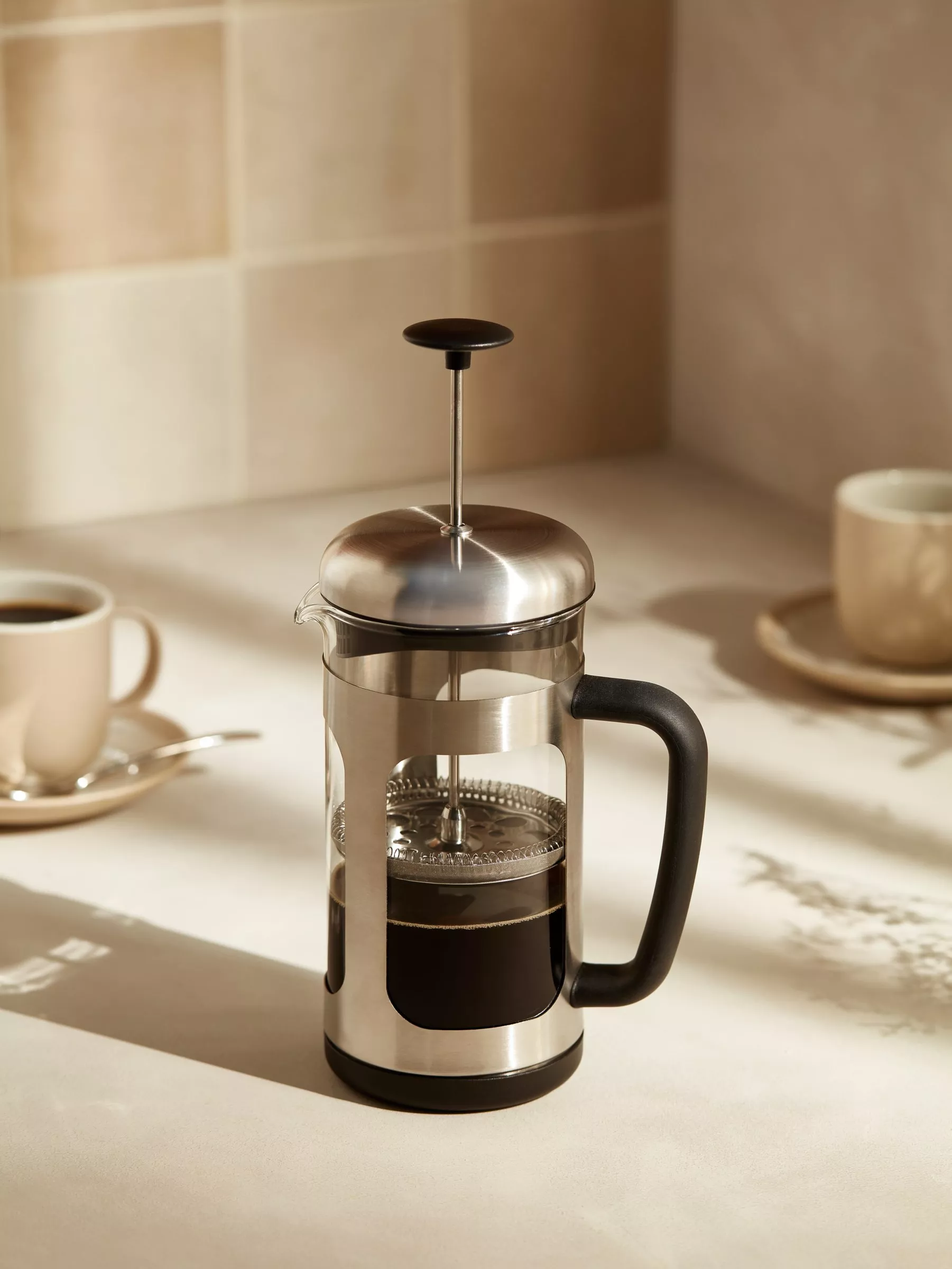 Stainless steel French press coffee maker on a kitchen worktop with mugs, brewing fresh coffee in sunlight