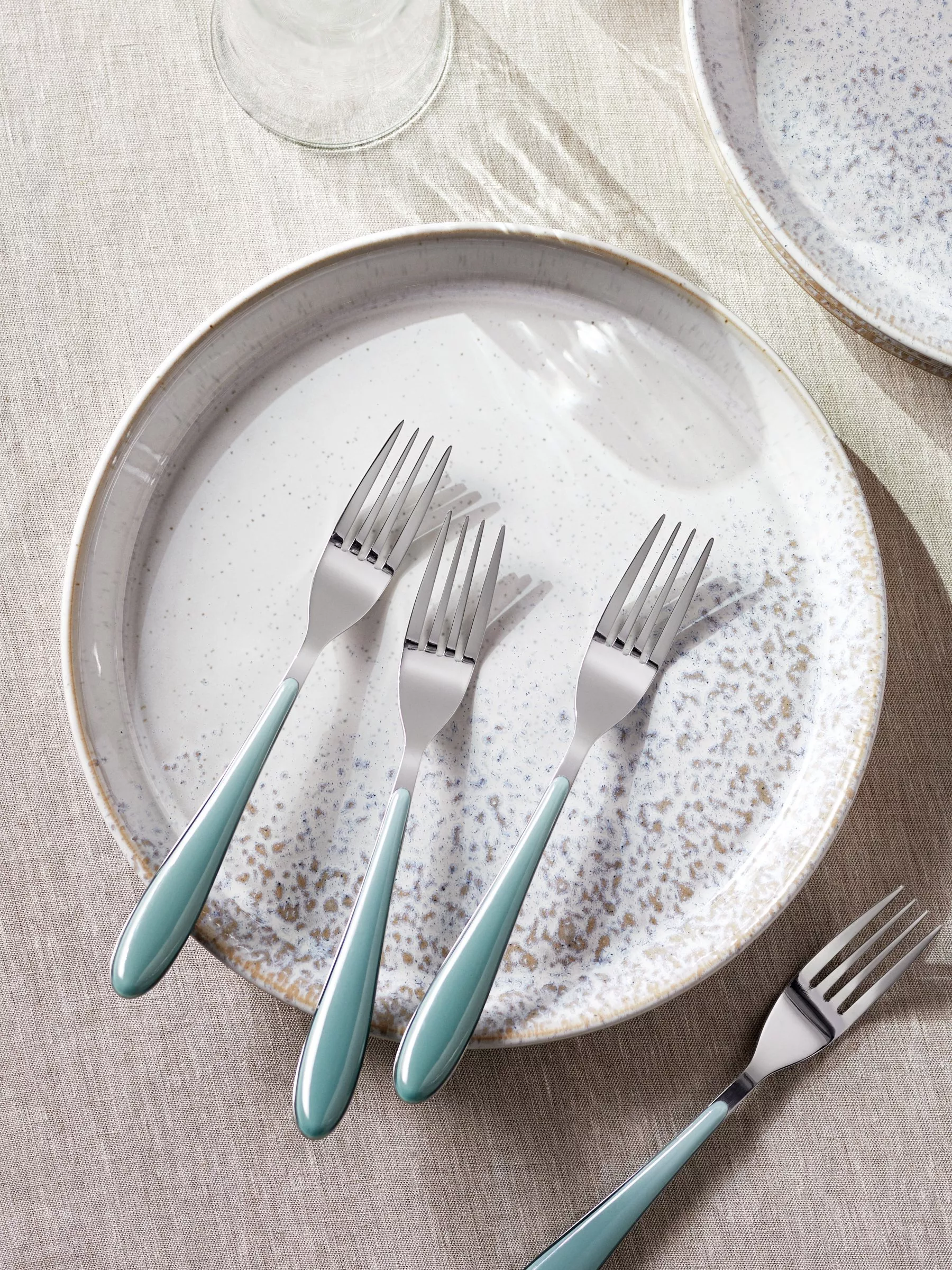 Three stainless steel dinner forks with teal handles on a speckled ceramic plate on a linen tablecloth