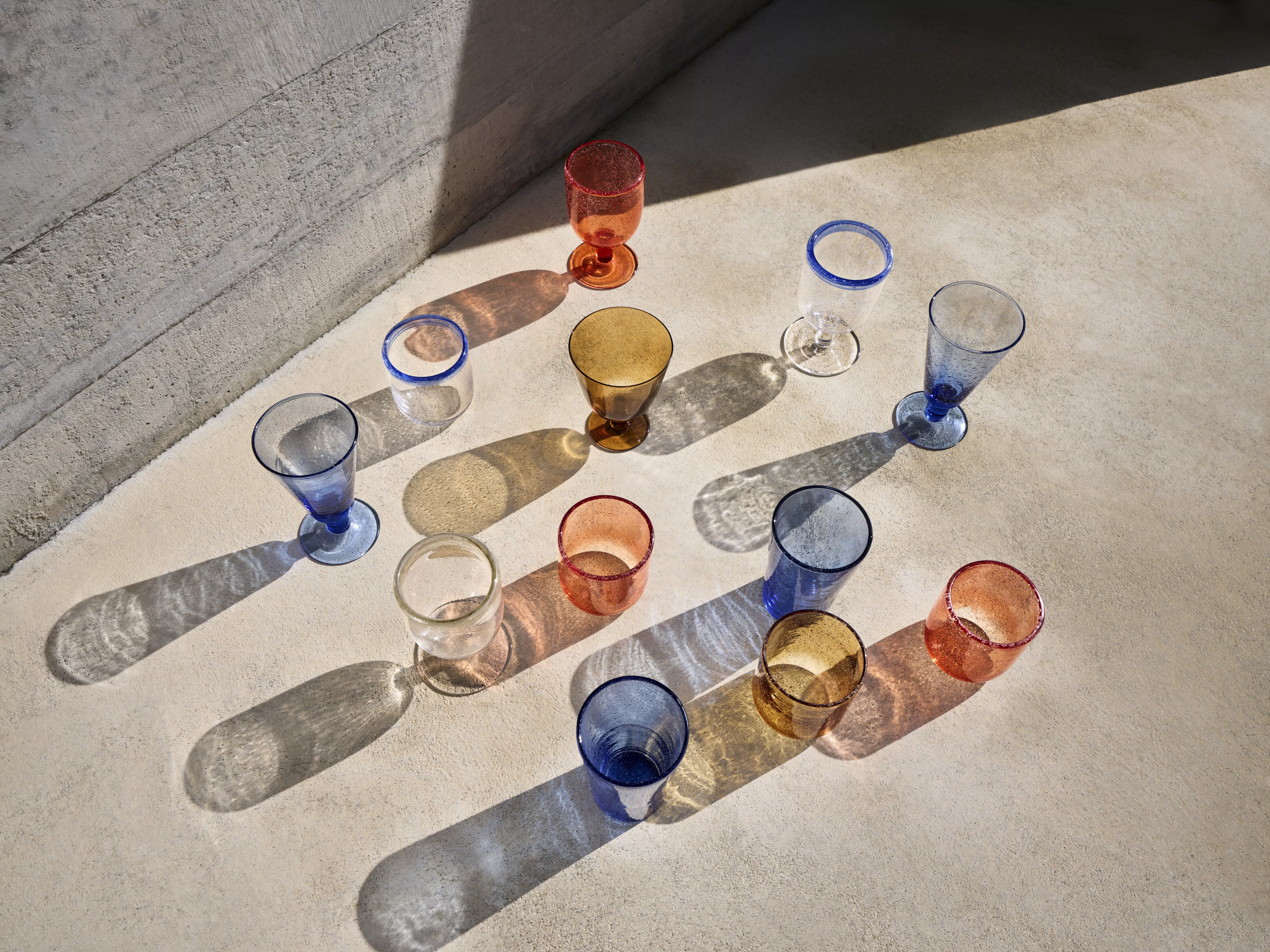 Colourful handmade glass tumblers and goblets on a sunlit concrete floor, casting long patterned shadows.