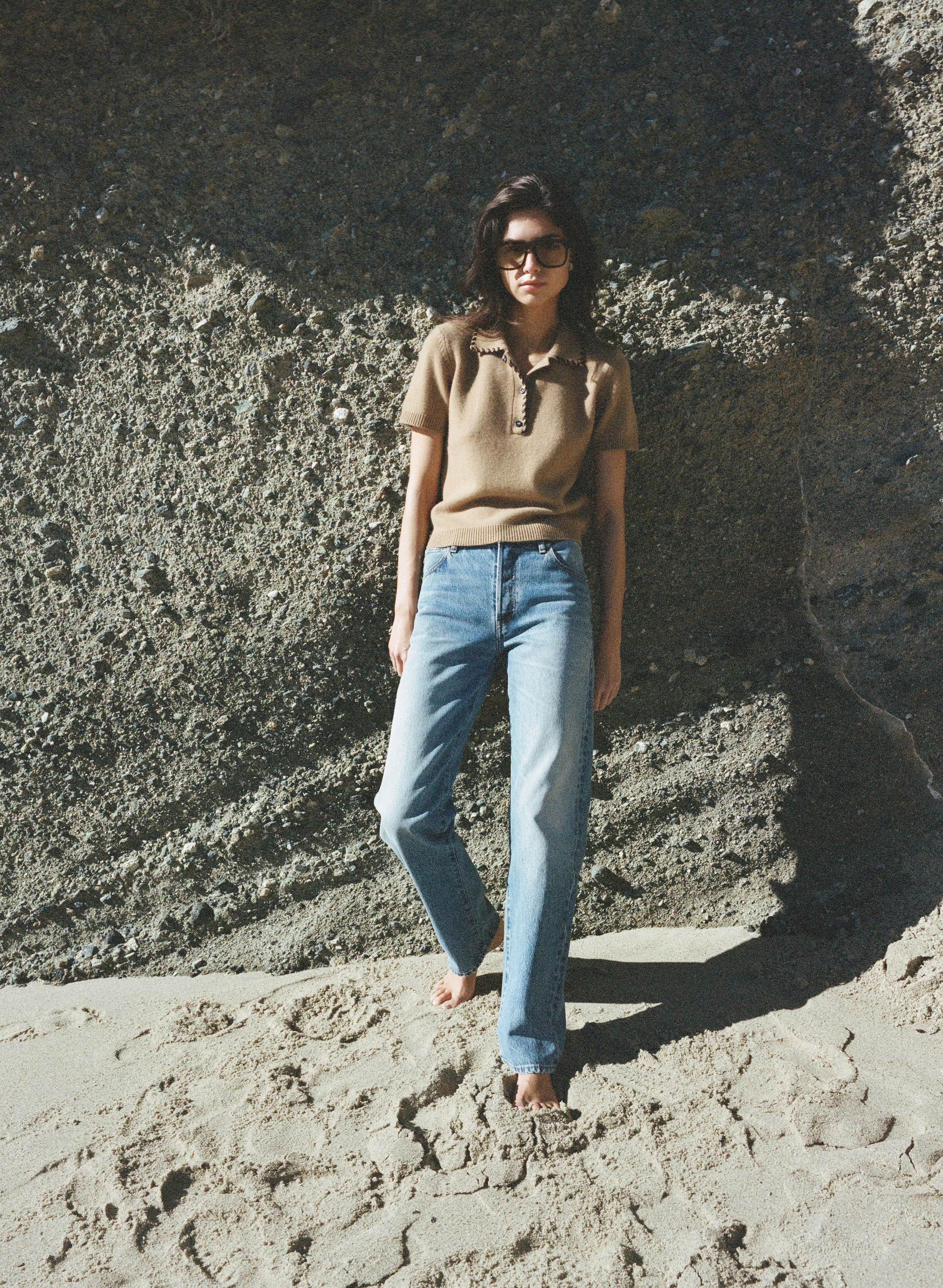 A person wearing a brown polo shirt and blue jeans stands barefoot on a sandy beach against a rocky background.
