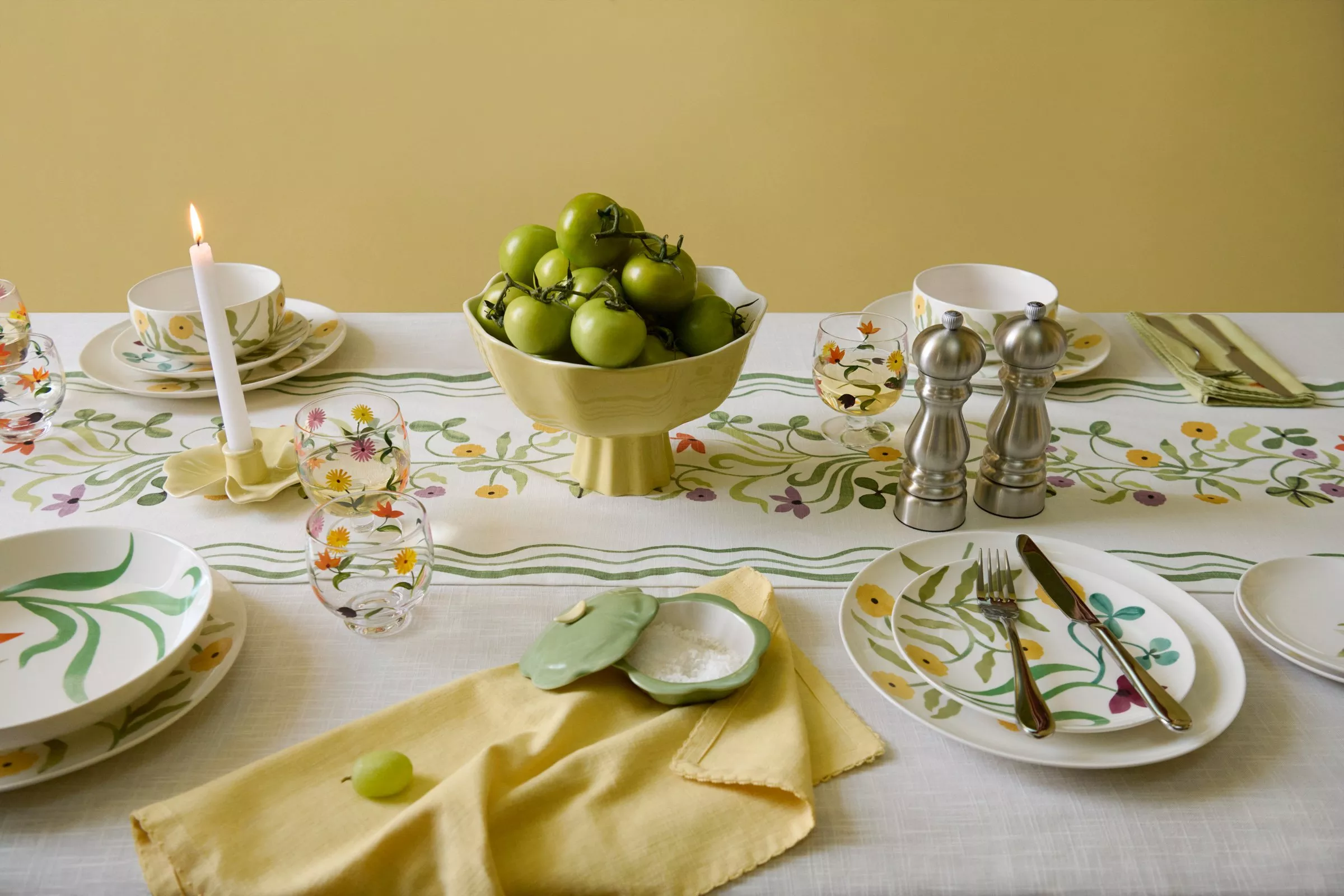 Elegant table setting with floral designs, featuring green tomatoes, candlelight, and silver salt and pepper shakers.