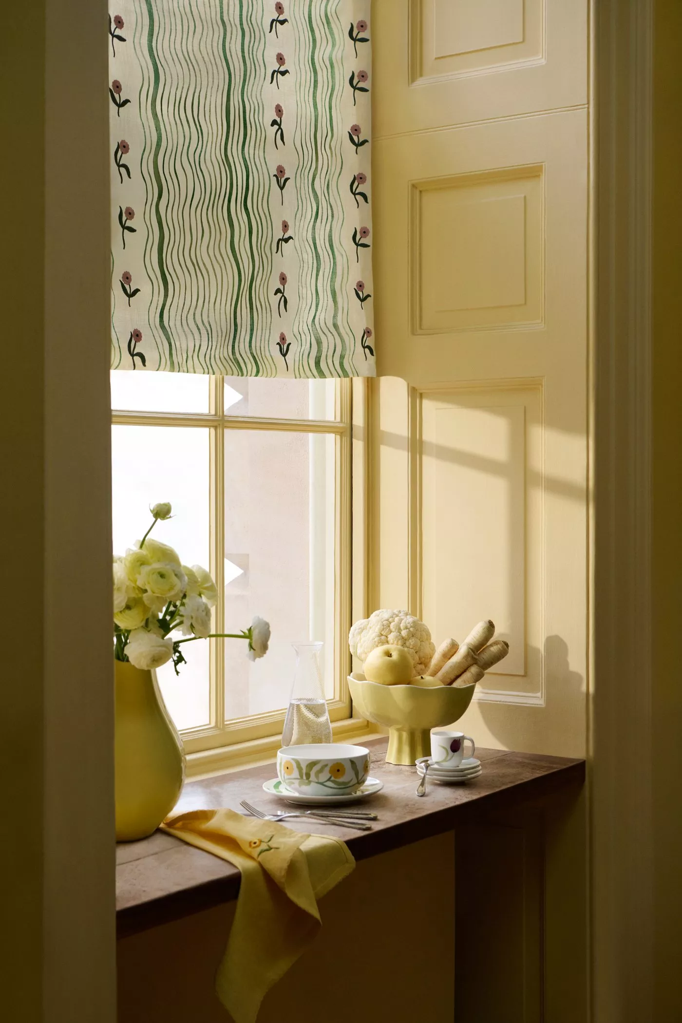 Sunny kitchen window with floral blind, yellow vase with white flowers, bowl of vegetables, and tableware.