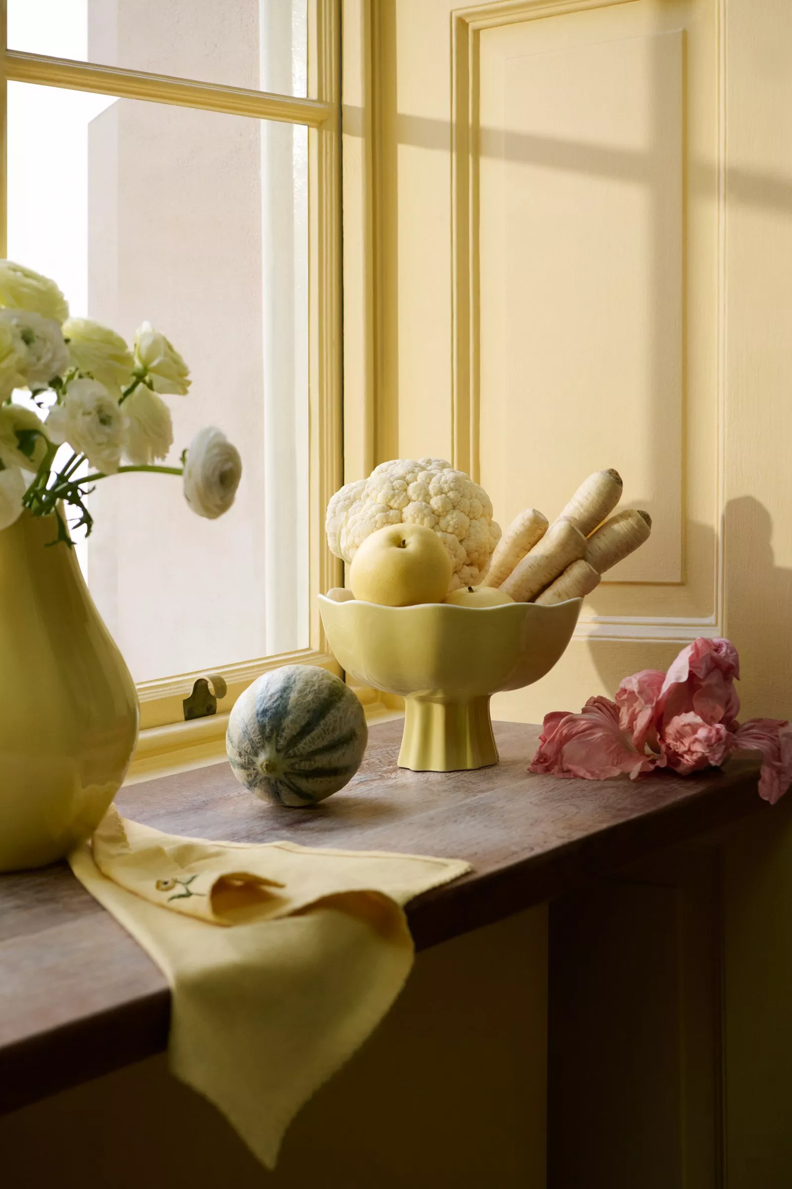 Sunny kitchen window scene with a vase of white flowers, fruits, and vegetables in a yellow bowl.