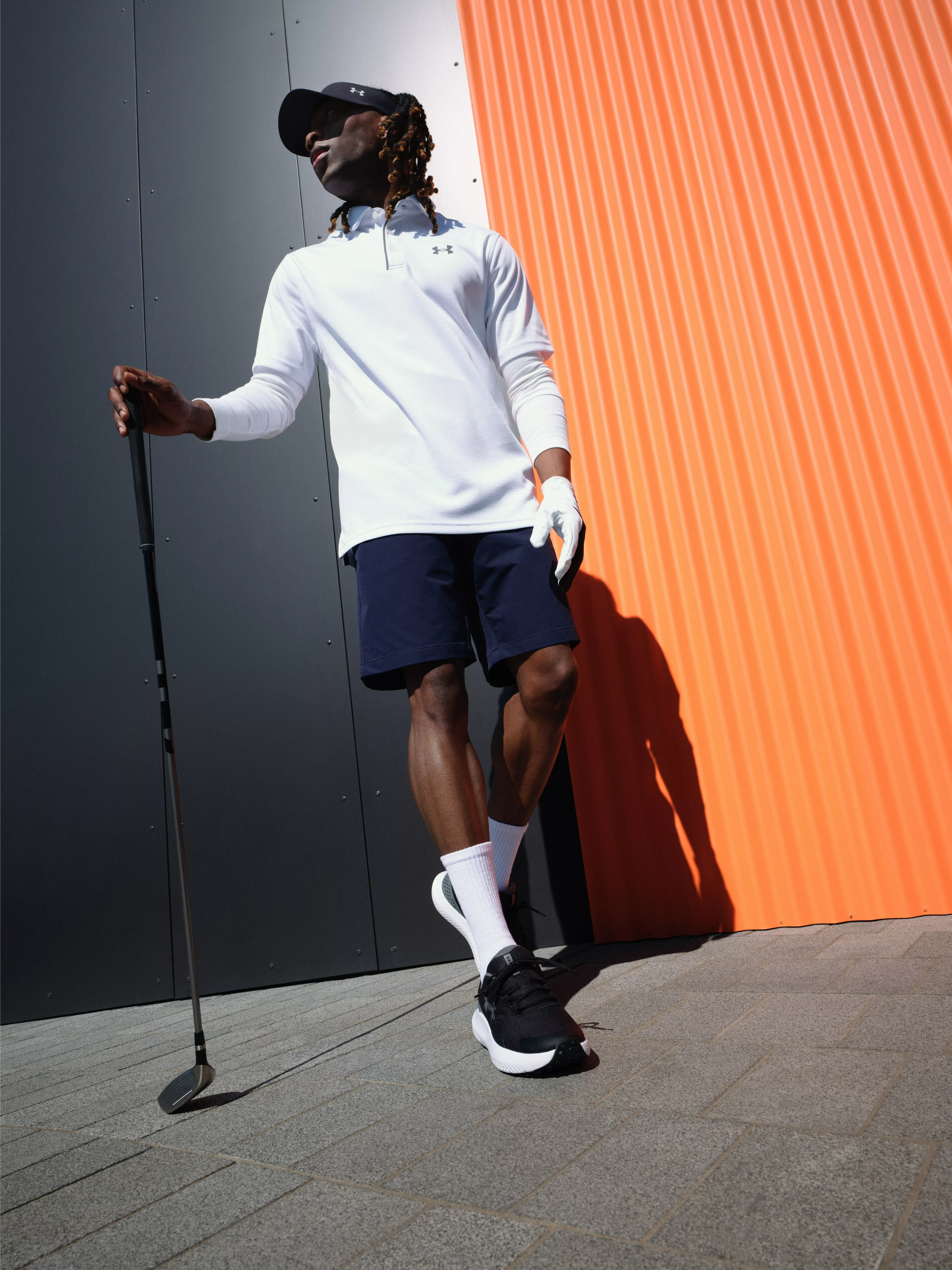 Golfer in white polo and navy shorts holding a club, leaning by orange and grey wall outdoors in sunlight.