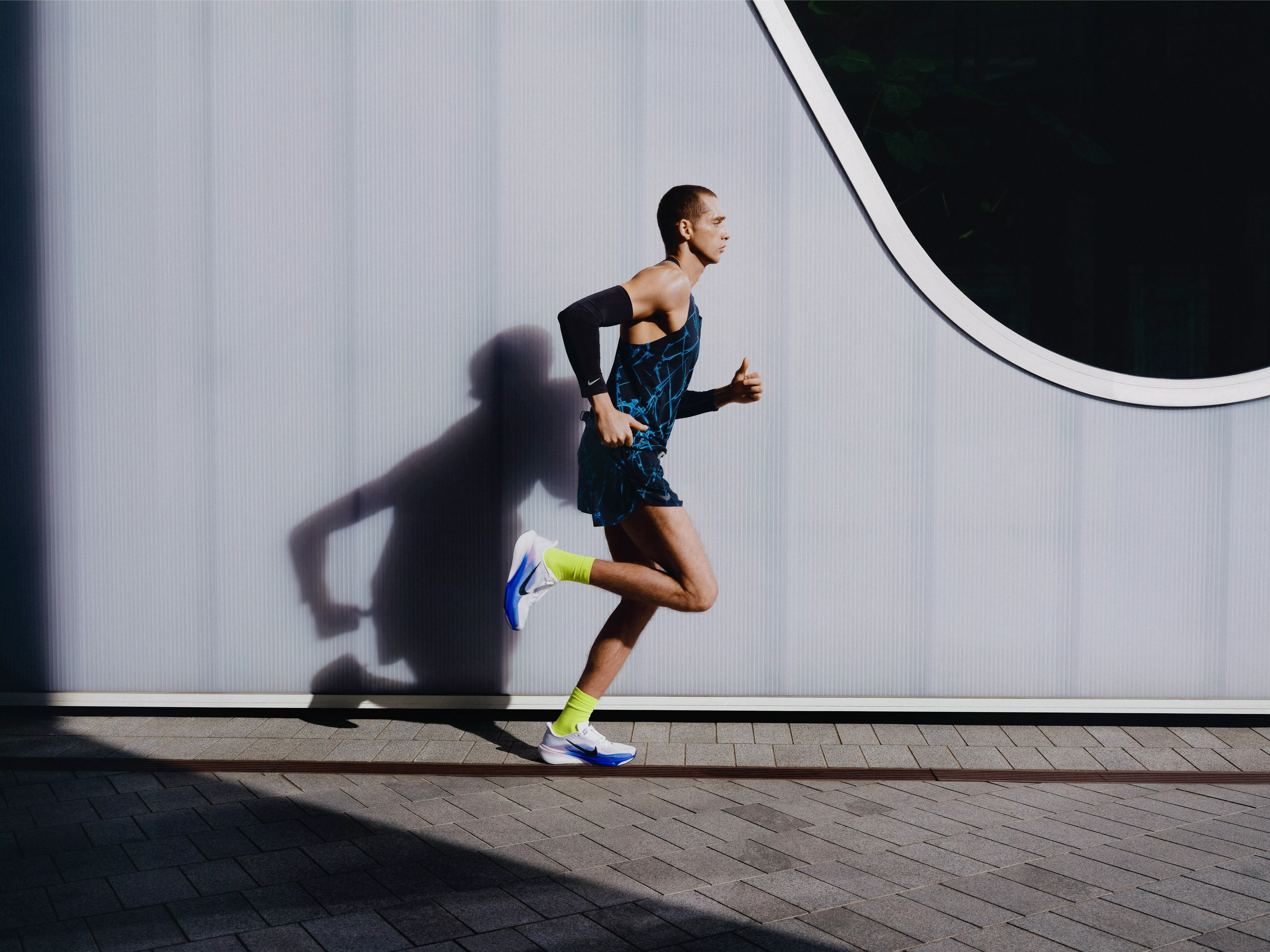 Male runner sprinting past modern building wall, wearing neon socks and trainers, casting sharp shadow outdoors