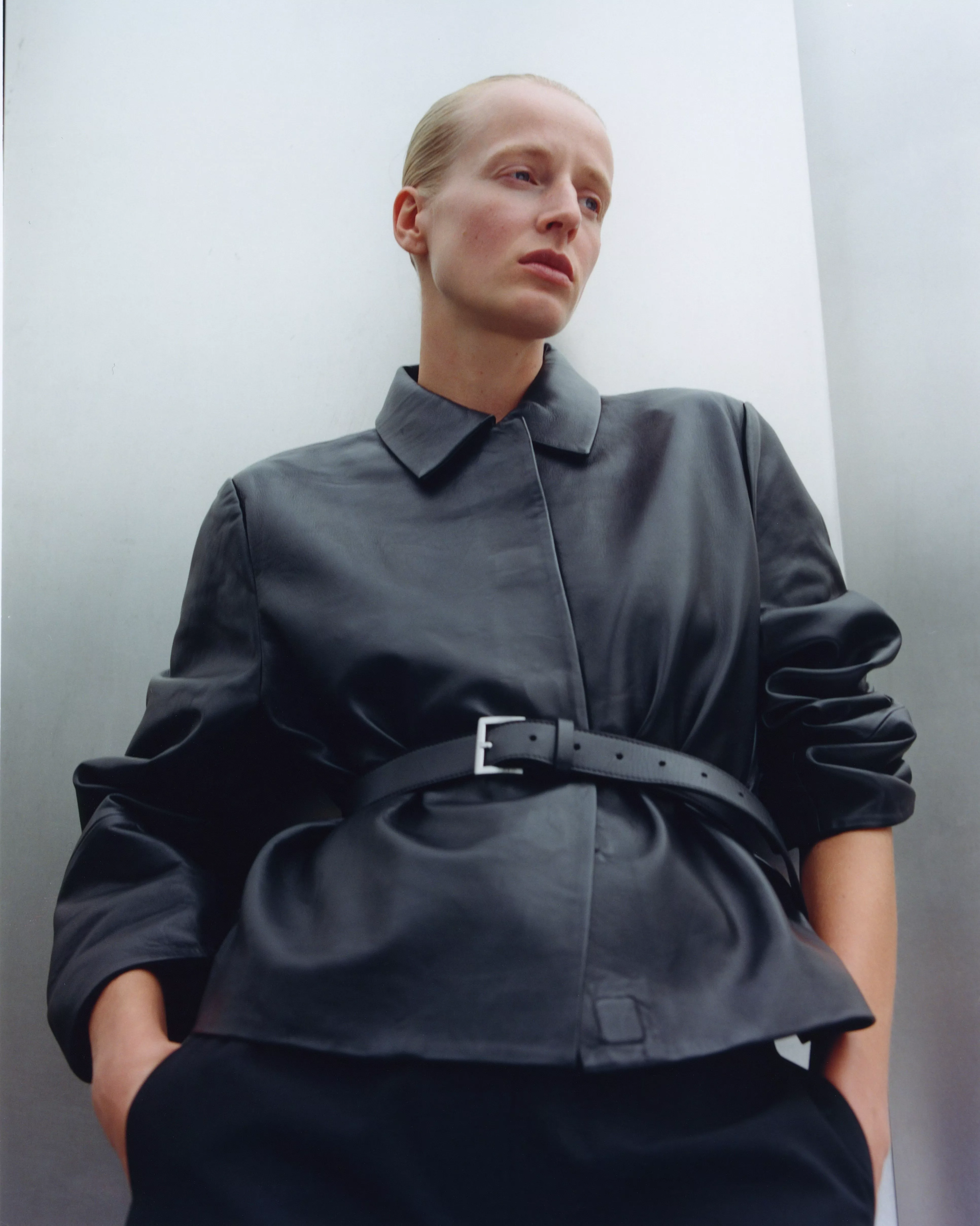 Fashion portrait of a model in a dark leather coat with belt, minimalist styling, looking away outdoors