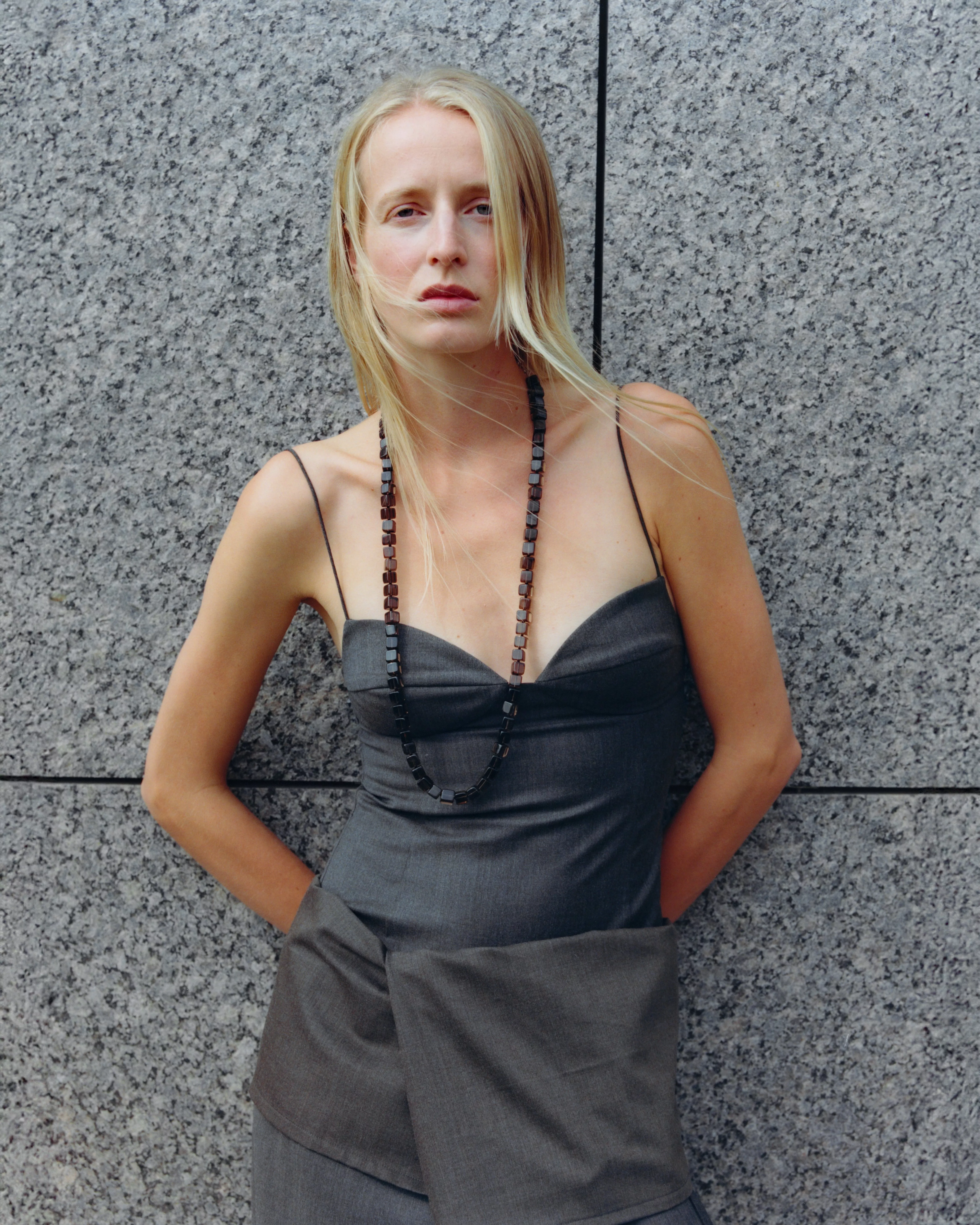 Blonde woman in a grey strapless dress with necklace, posing against a granite wall outdoors in daylight