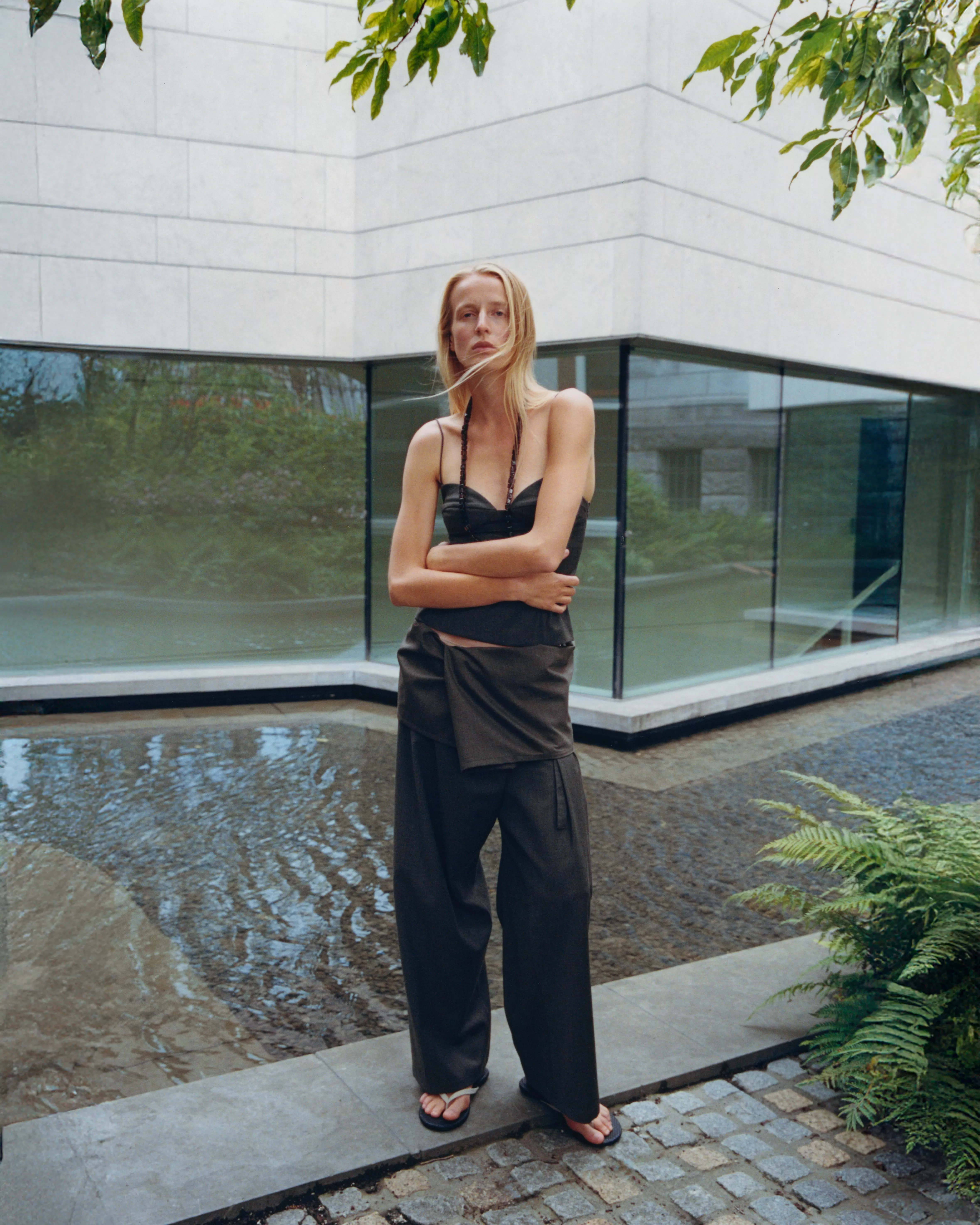 Fashion model in black corset top and wide-leg trousers by modern glass building beside shallow water feature