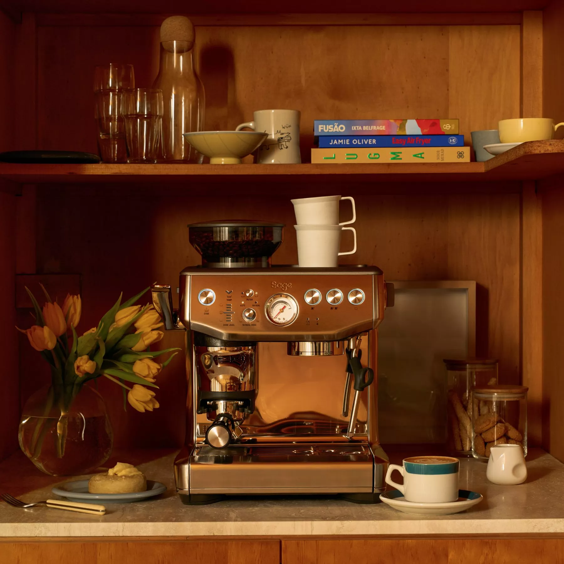 Stainless steel espresso machine on kitchen counter with coffee cups, cookbooks, glassware and yellow tulips.