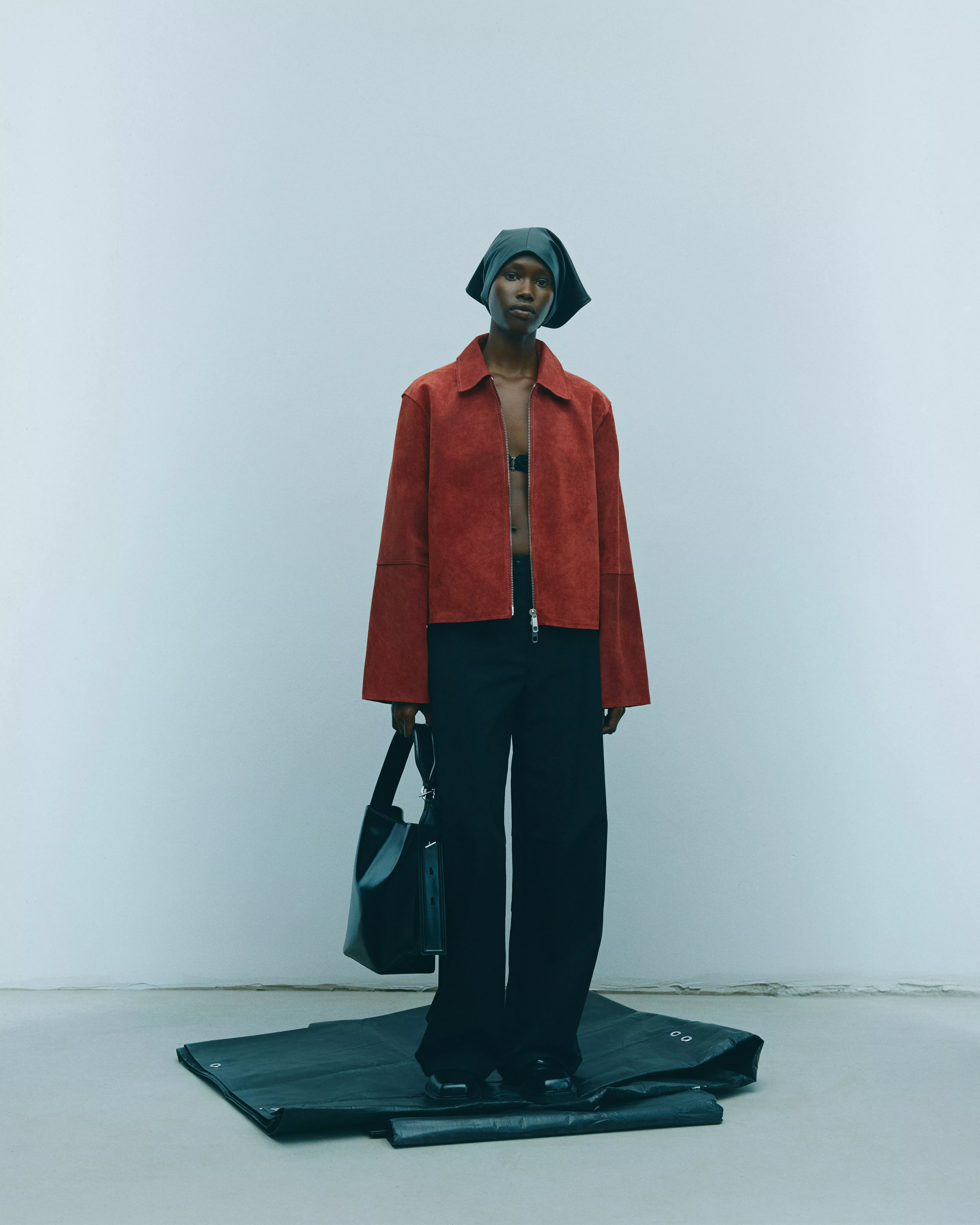 Fashion model wearing a red suede jacket and wide-leg black trousers, holding a tote bag in minimalist studio setting