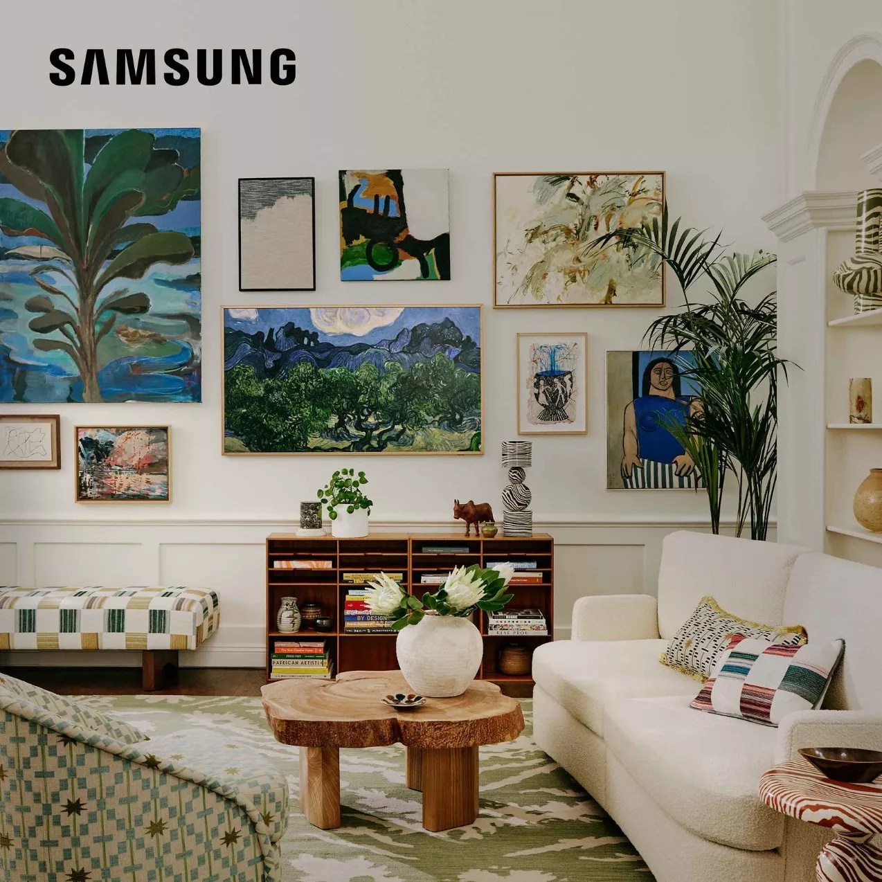 A Samsung Frame TV displaying a classic landscape artwork blends into a gallery wall in a stylish living room with a white sofa, patterned armchairs, and a wooden coffee table.