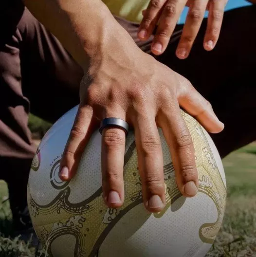 Close-up of a hand wearing a black silicone ring gripping a rugby ball on grass, outdoor sports action shot