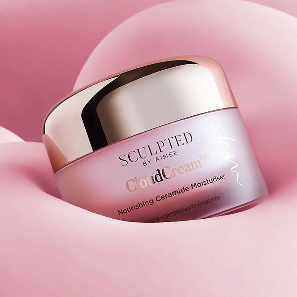 A luxurious pink and gold jar of Sculpted by Aimee CloudCream Nourishing Ceramide Moisturiser is displayed against a soft, cloud-like pink background.
