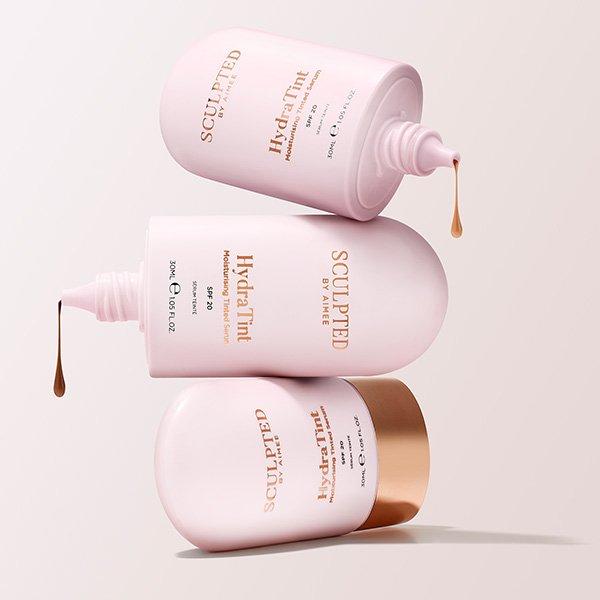 Three pink bottles of Sculpted by Aimee HydraTint moisturising skin tint with SPF 20 are shown stacked and dripping product, highlighting their smooth, fluid texture.