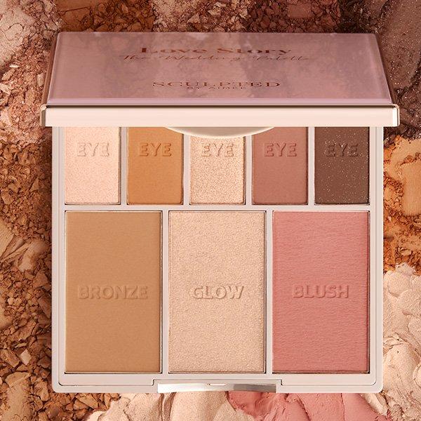 An open Sculpted by Aimee makeup palette displays five eye shadows, a bronzer, highlighter labeled “Glow,” and a blush, set against a textured powder background.