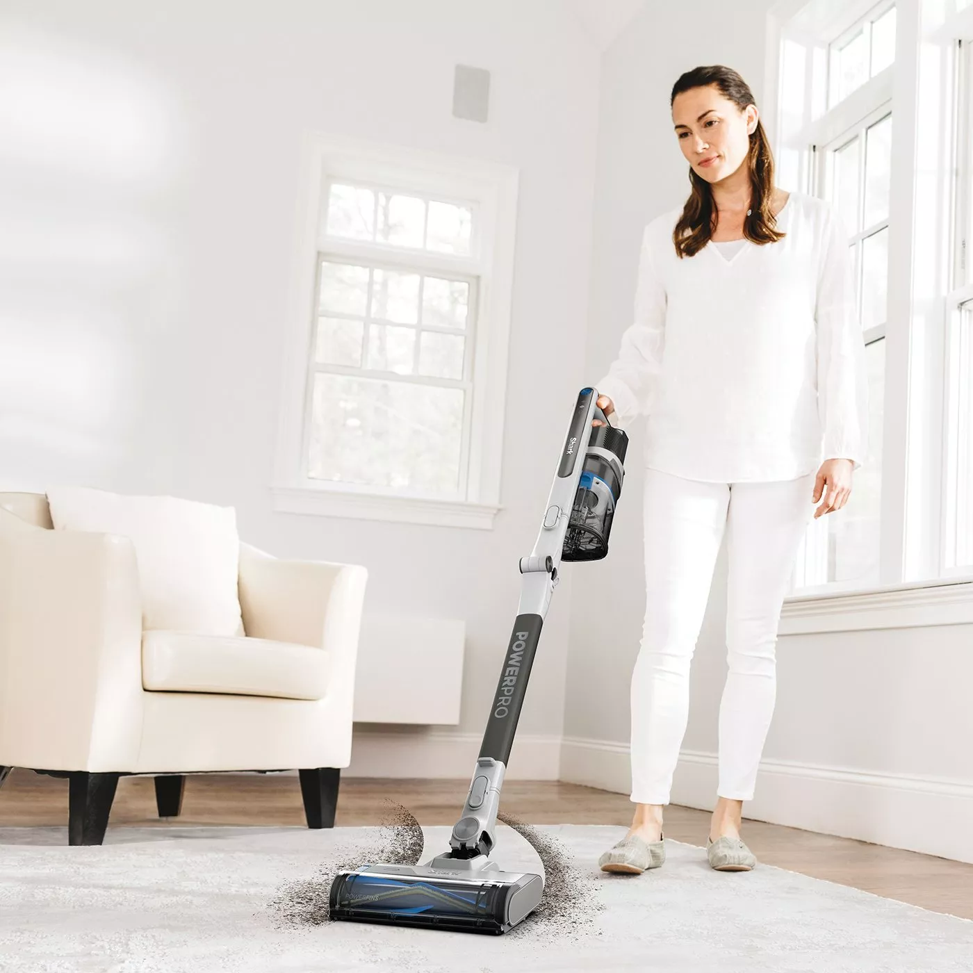 Woman using cordless stick vacuum cleaner on living room rug in bright modern home interior near sofa