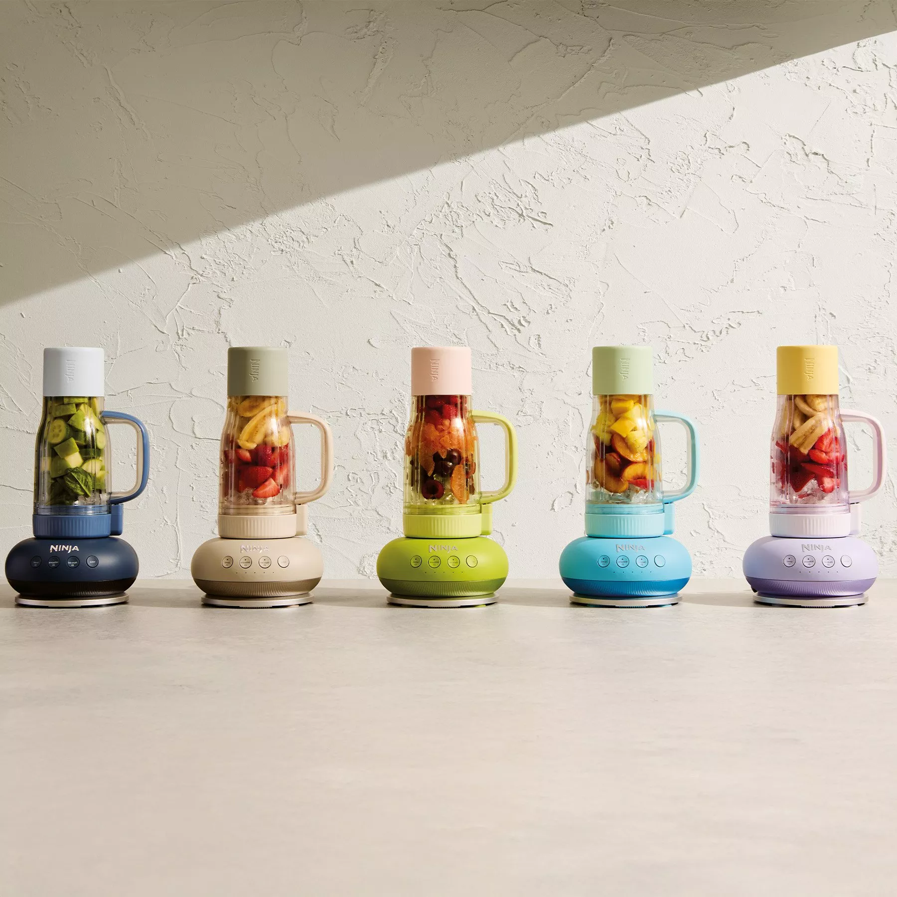 A row of colourful blenders filled with fruit, set against a textured wall. Each blender features a different pastel hue.