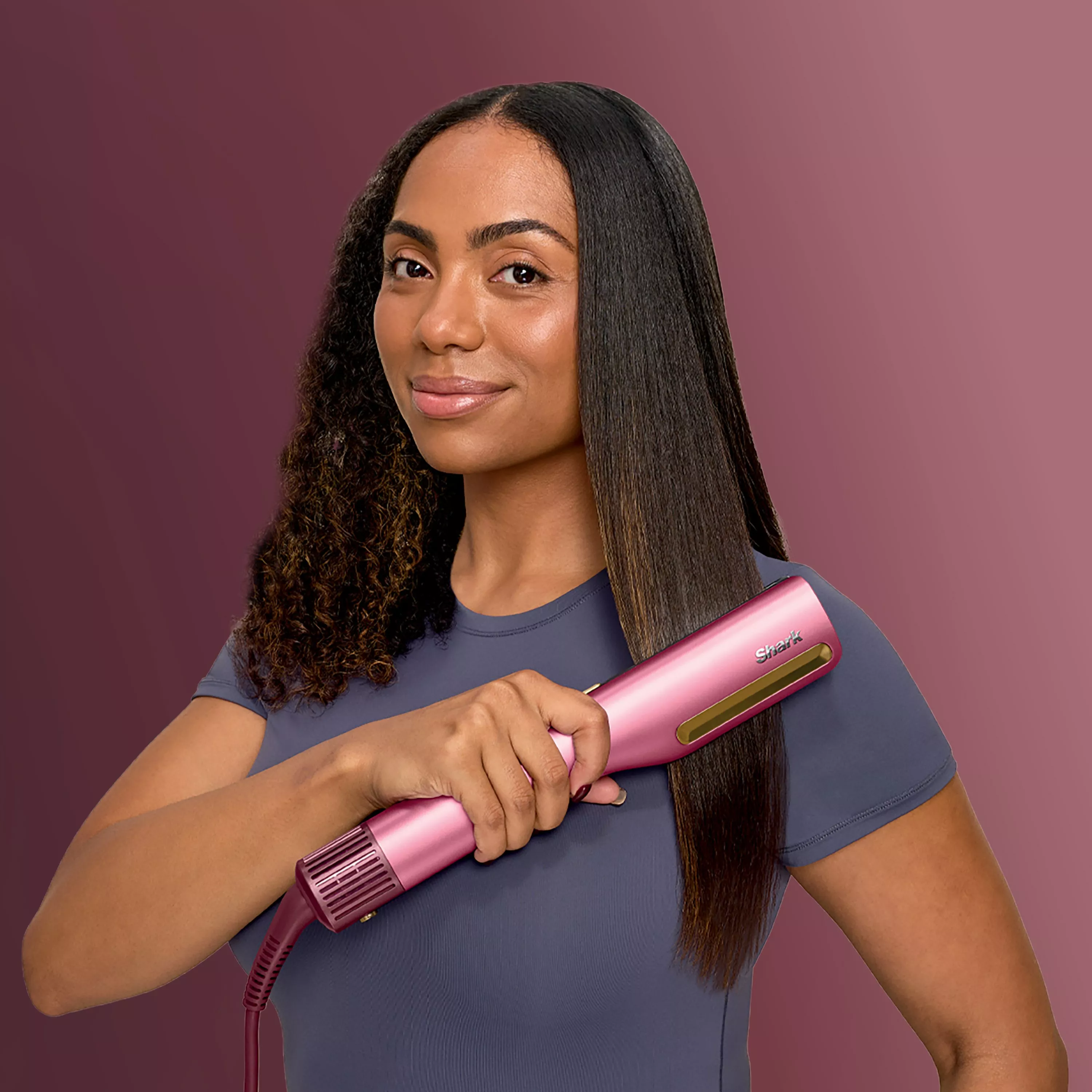A person using a pink hair straightener, showcasing half curly and half straight hair on a purple background.