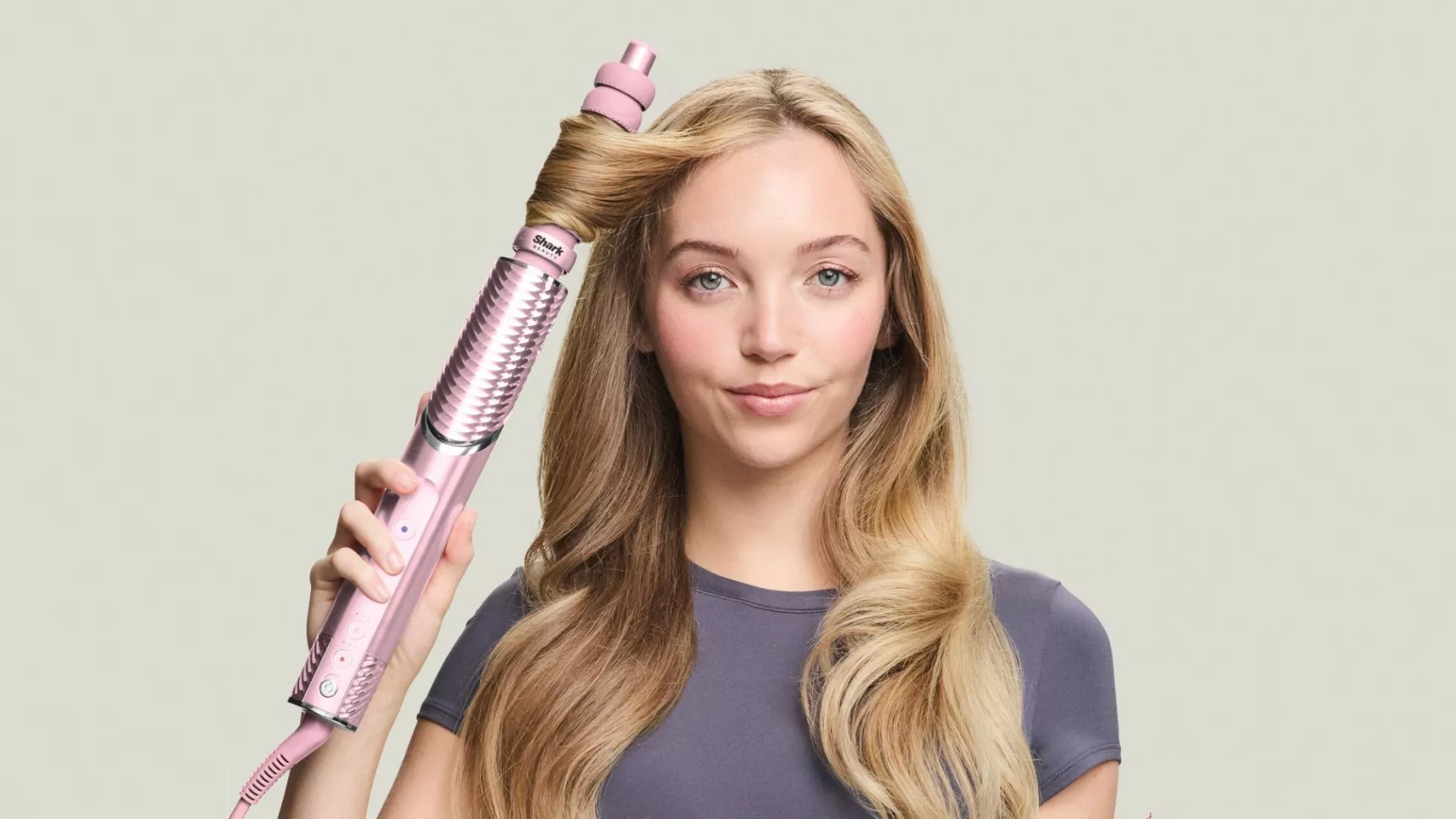 A blonde woman styles her long hair with a metallic pink Shark Beauty hair curler, wrapping a strand of hair around the barrel while looking directly at the camera against a plain gray background.