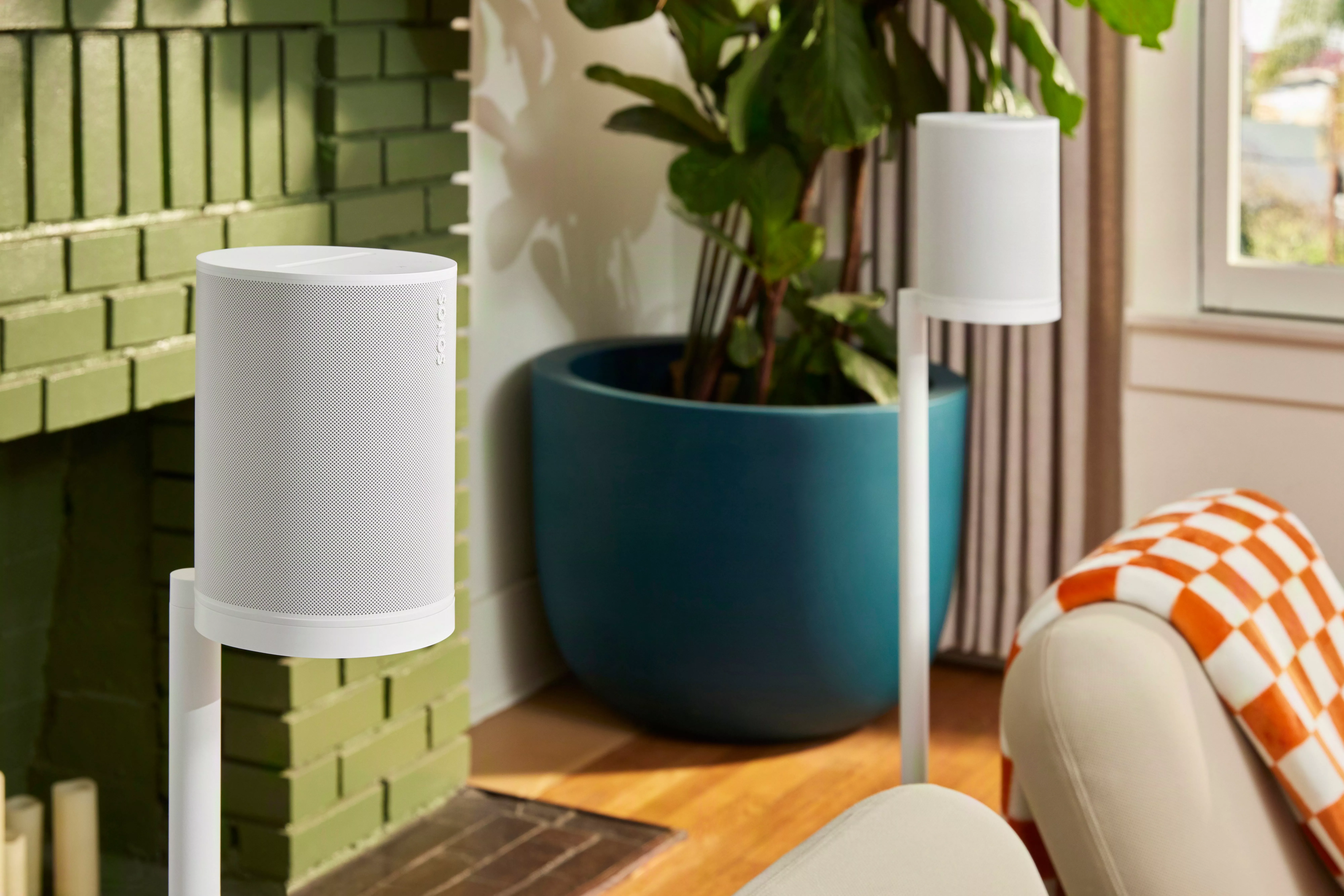 Modern white wireless smart speakers on stands in a cosy living room beside a green brick fireplace and plant