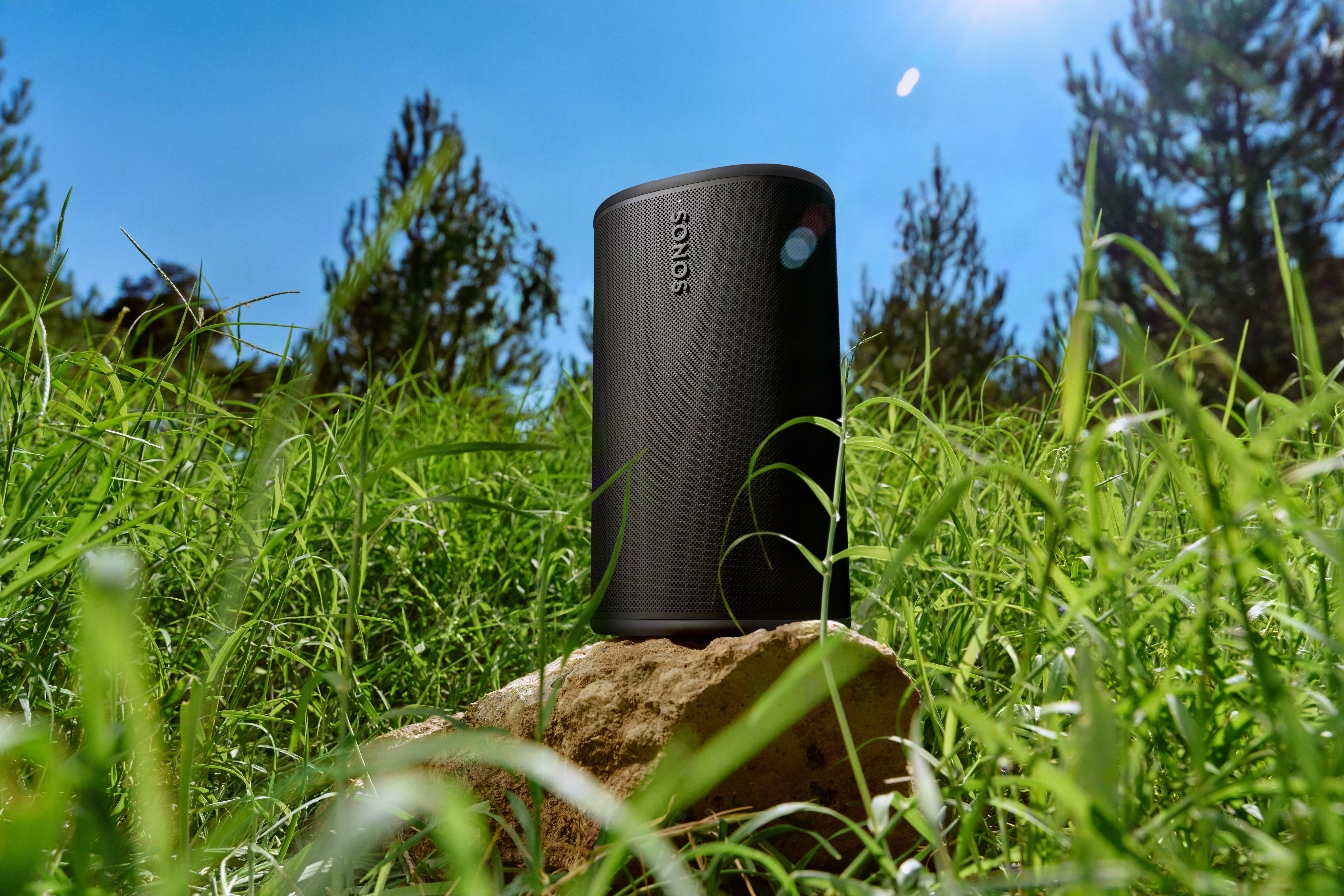 A portable black speaker on a rock amidst lush green grass, under a sunny blue sky, with trees in the background.