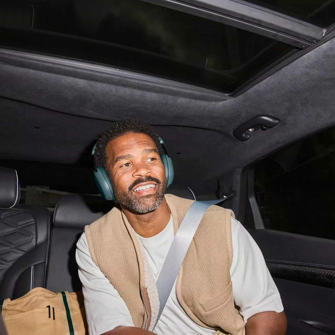 A man wearing dark green over-ear headphones and a tan vest sitting in the backseat of a car with a sunroof open.