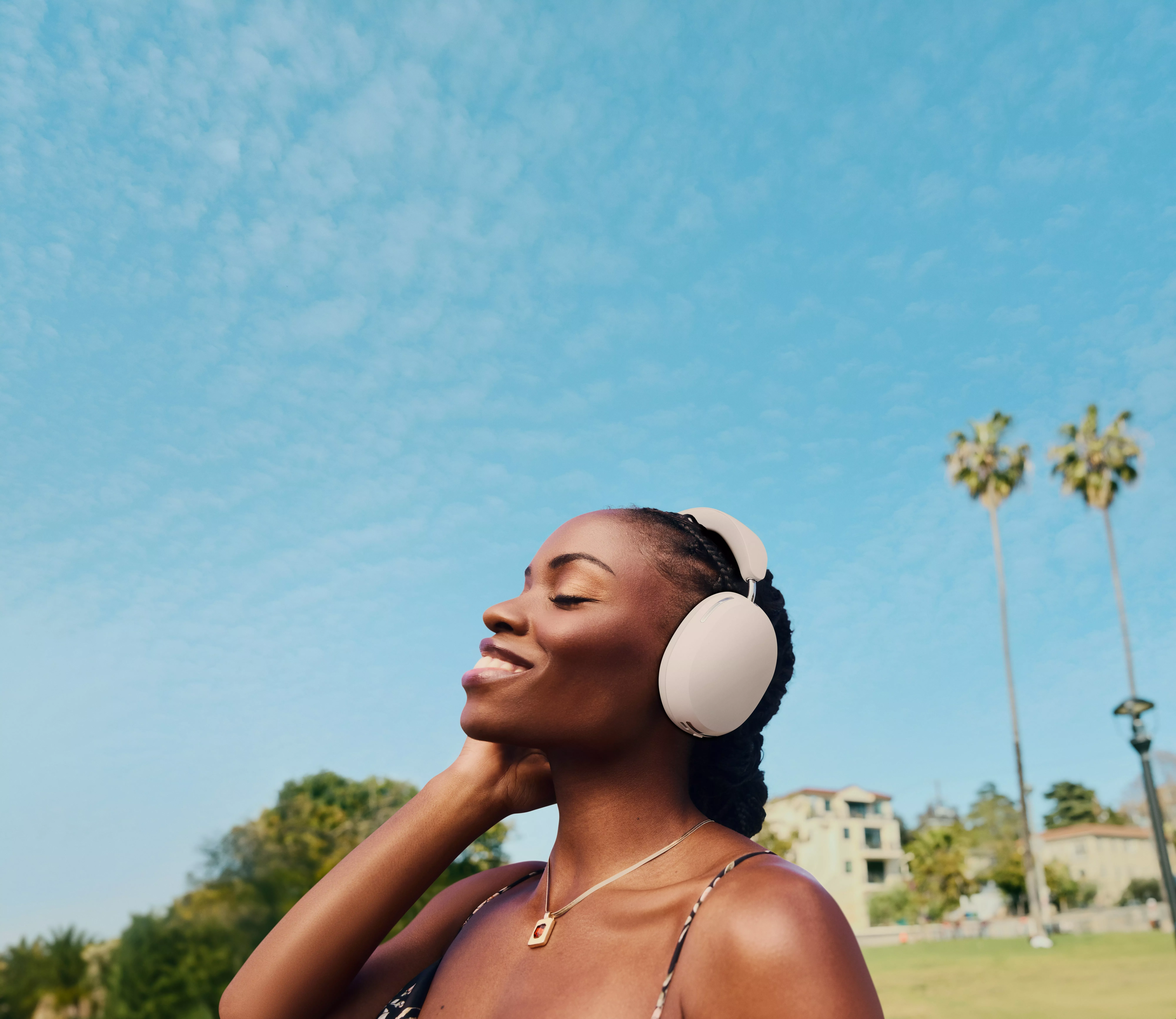 Smiling woman wearing wireless over-ear headphones outdoors under blue sky, enjoying music in a sunny park
